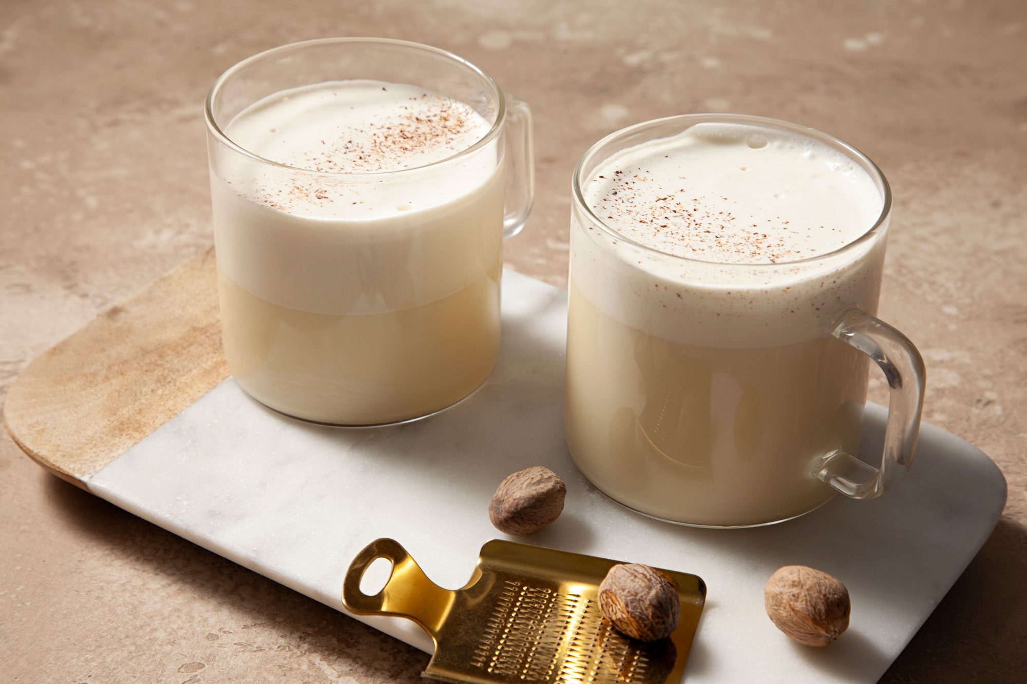 3/4 angle view shot of Homemade Eggnog; serve in two glasses on white tray; sprinkle with additional nutmeg before serving; light brown marble background;
