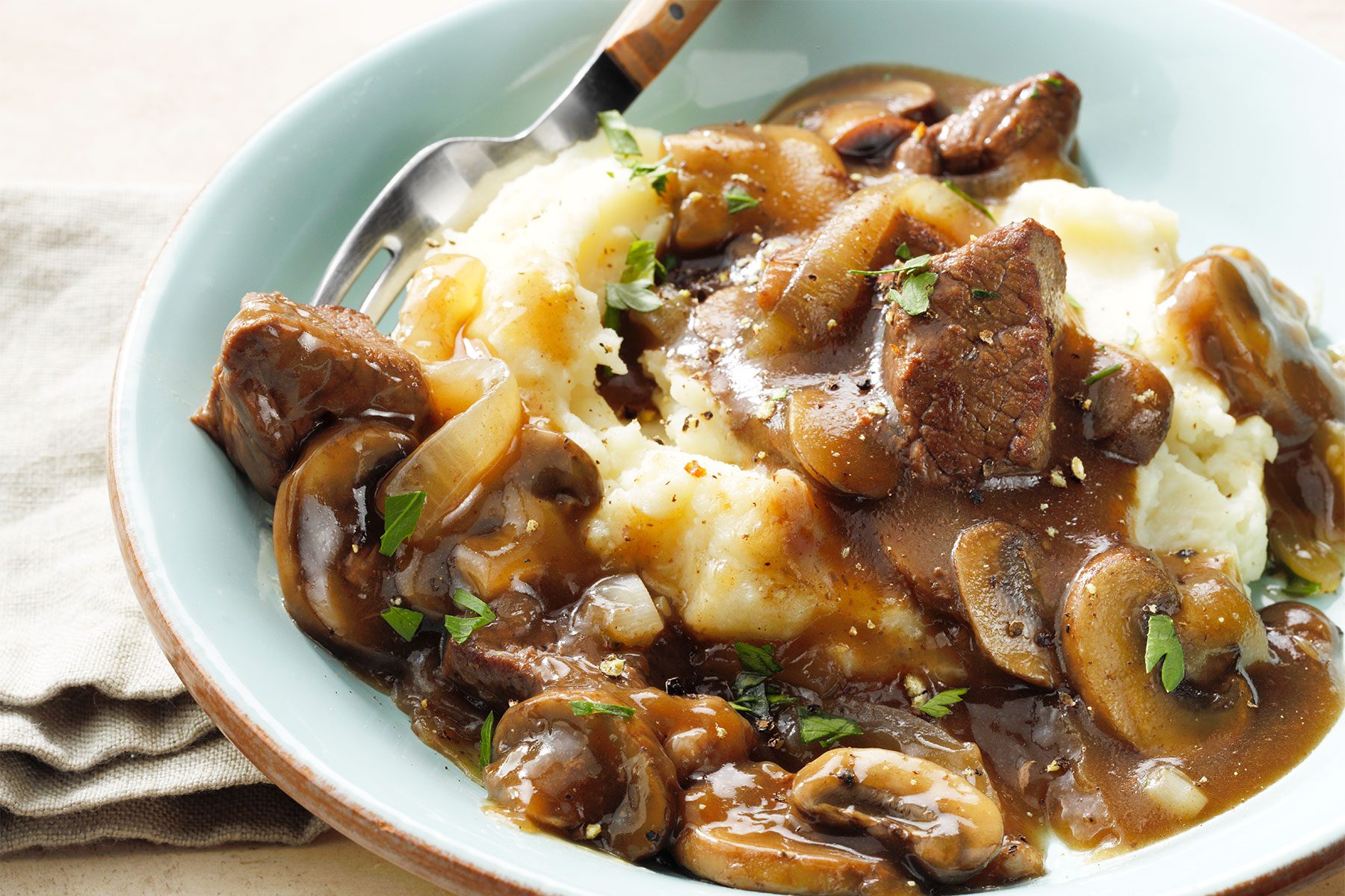 A plate of mashed potatoes topped with beef stew and mushrooms in a rich brown gravy. Parsley is sprinkled on top as a garnish. A fork rests on the edge of the plate, and a napkin is partially visible underneath.