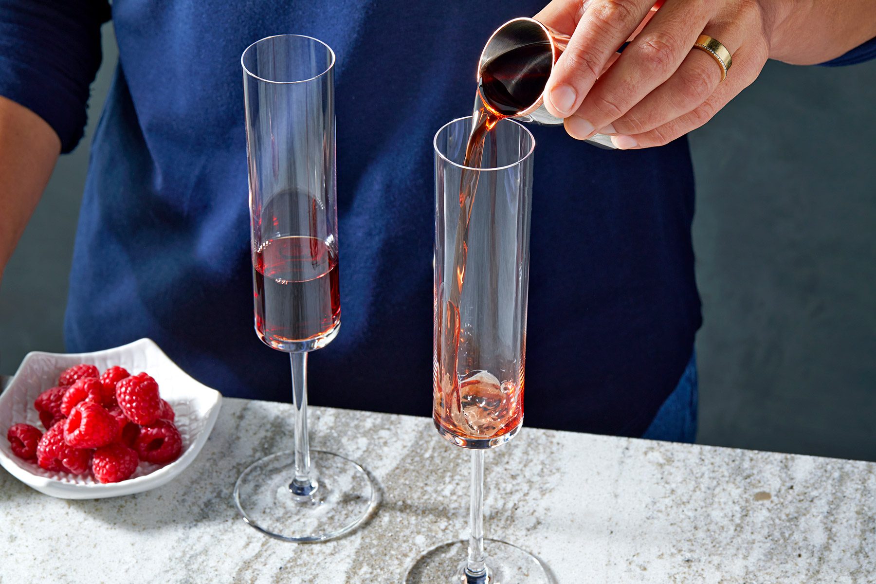 3/4 angle view shot of pour crème de cassis into champagne flute glass; bowl of raspberries; marble backgroundl;