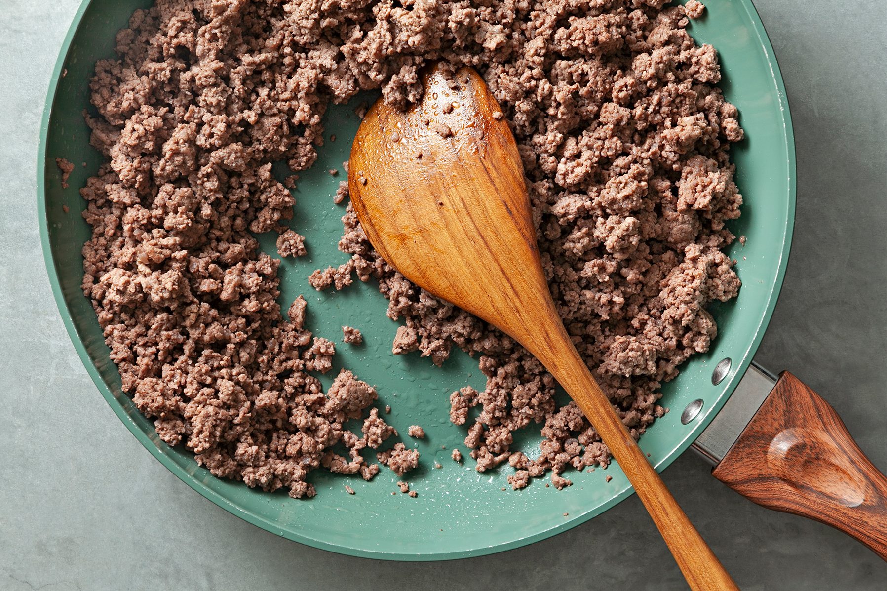 A green frying pan with a wooden handle is filled with browned ground beef. A wooden spoon rests in the center of the pan, positioned diagonally.