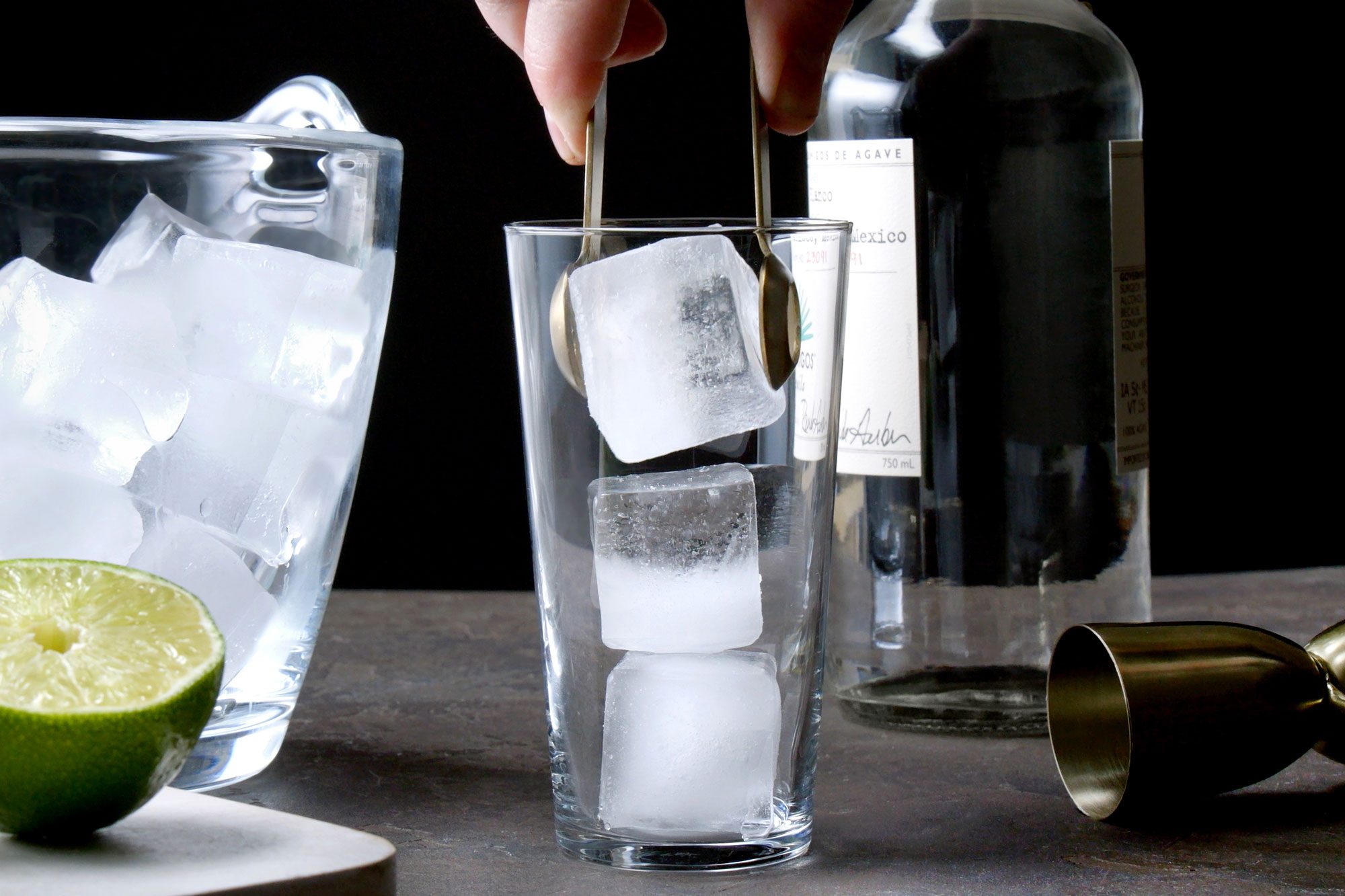 Table view shot of fill highball glass about two-thirds full with ice; half cut lime; chopping board; black texture background;