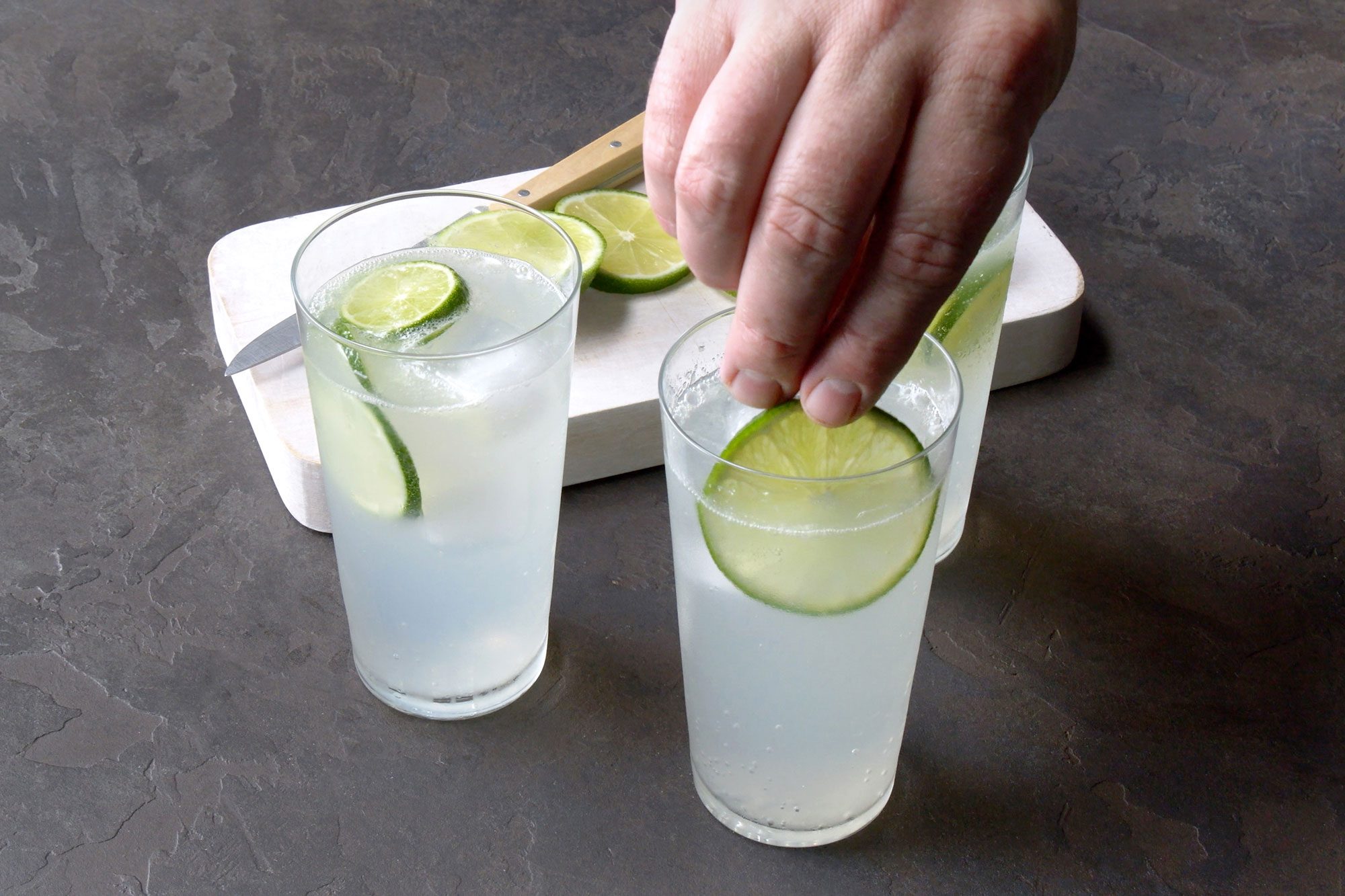 High angle view shot of ranch water in highball glasses garnished with lime slices; wooden chopping board; knife; black texture background;