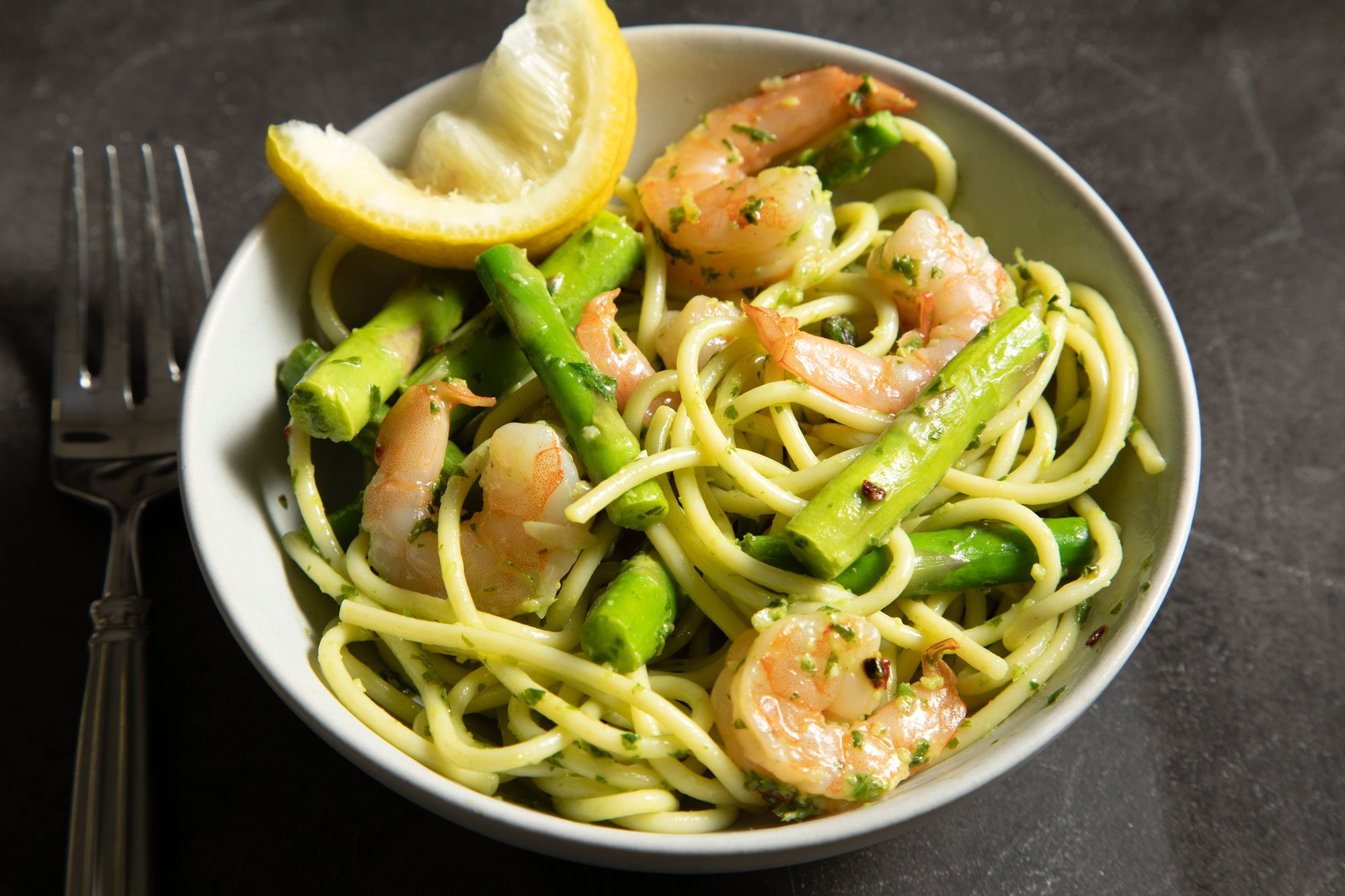 3/4th shot; black background; Pesto Shrimp Pasta served on white plate with silver fork; lemon wedge;