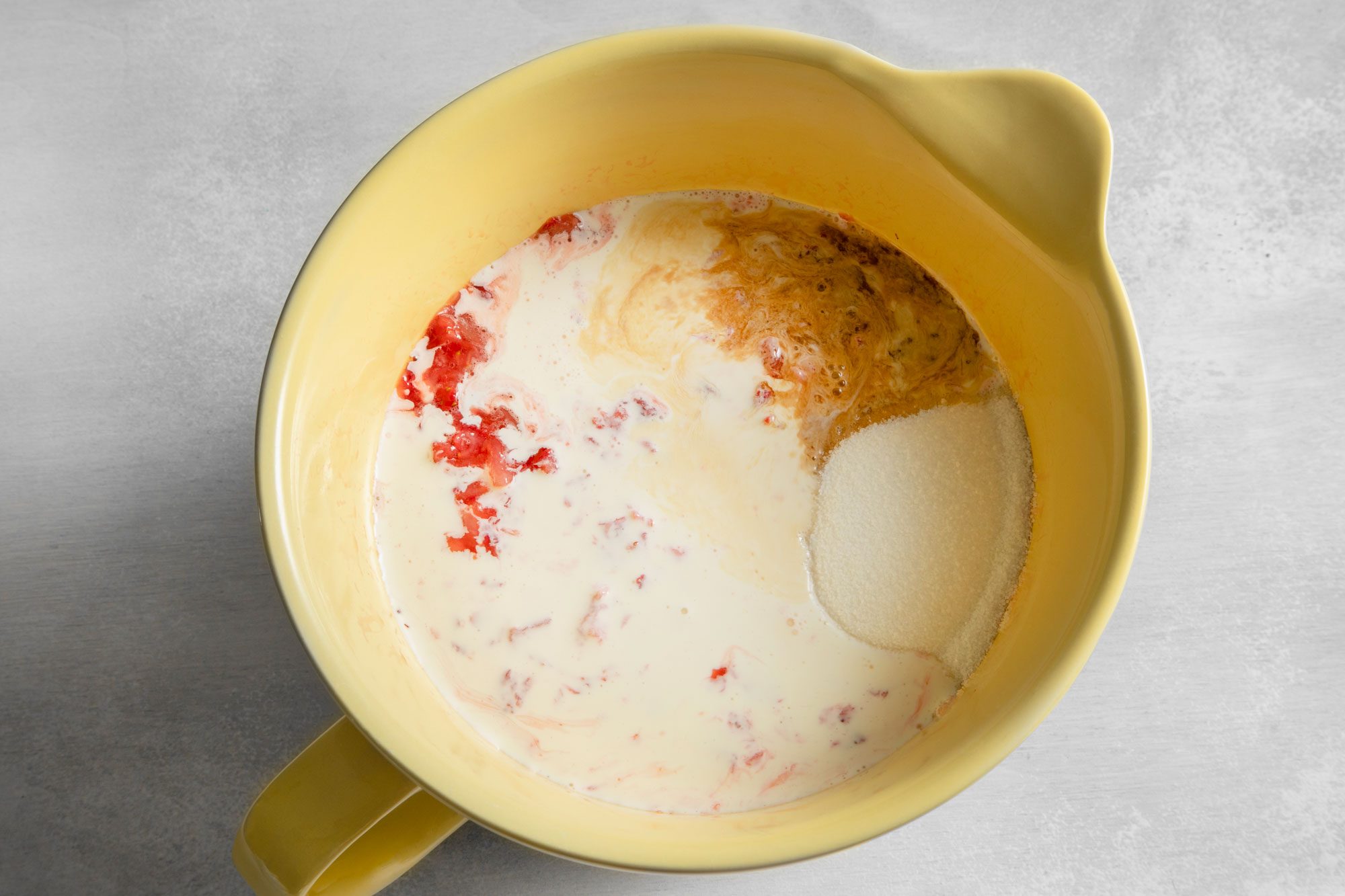 Overhead shot of milk, sugar, mashed strawberries and vanilla extract in large bowl on white marble background