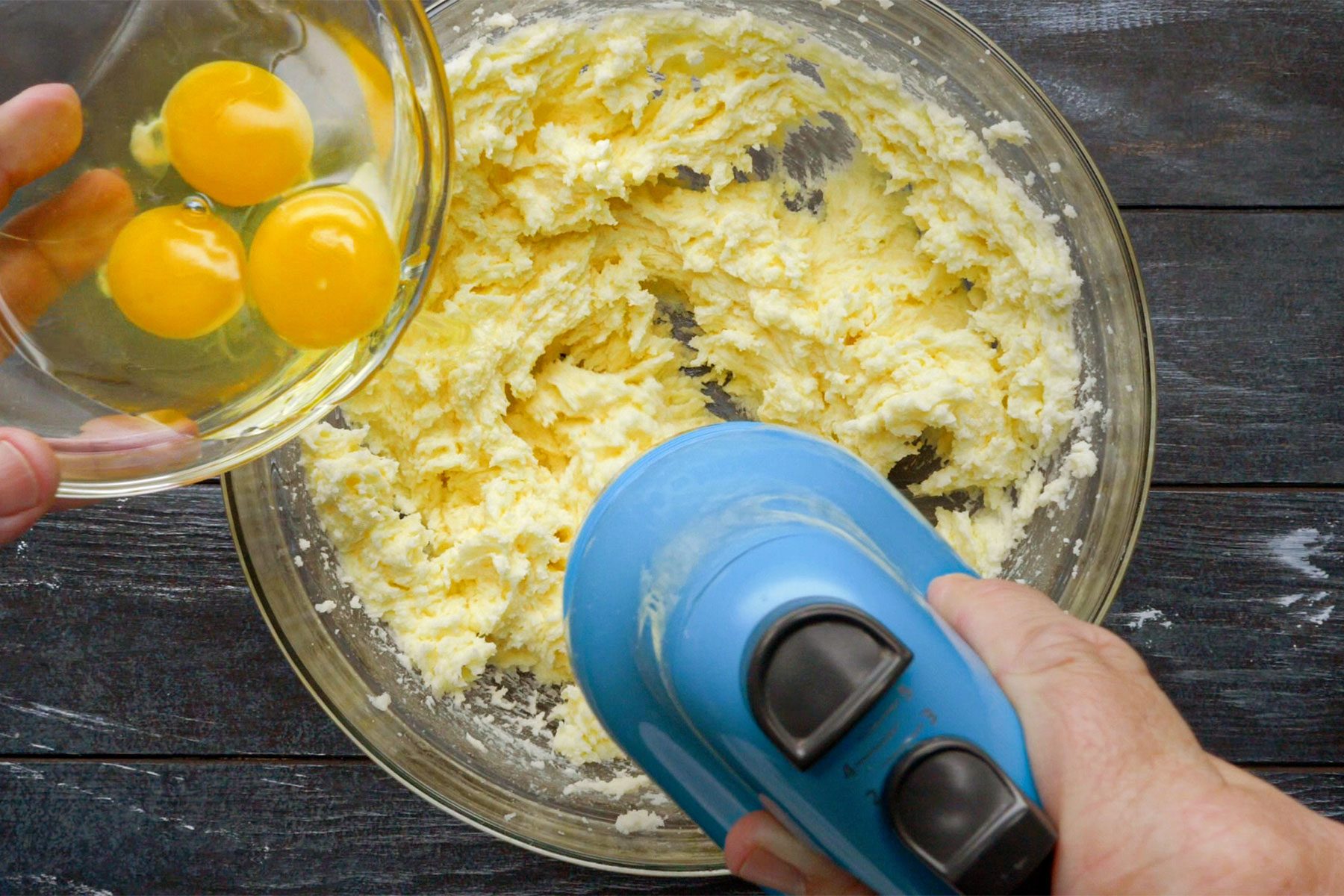 Overhead shot of large bowl; adding eggs; 1 at a time; beating well after each addition; dark wooden background;