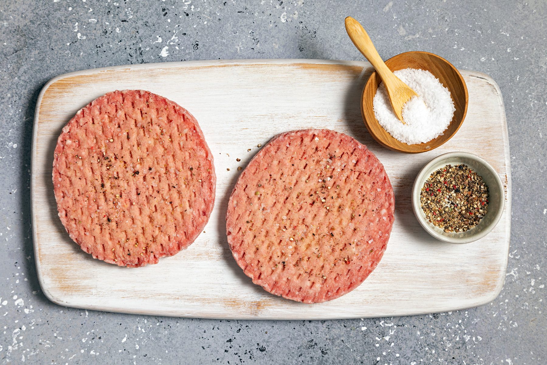 Two raw beef patties with salt and pepper on a wooden board