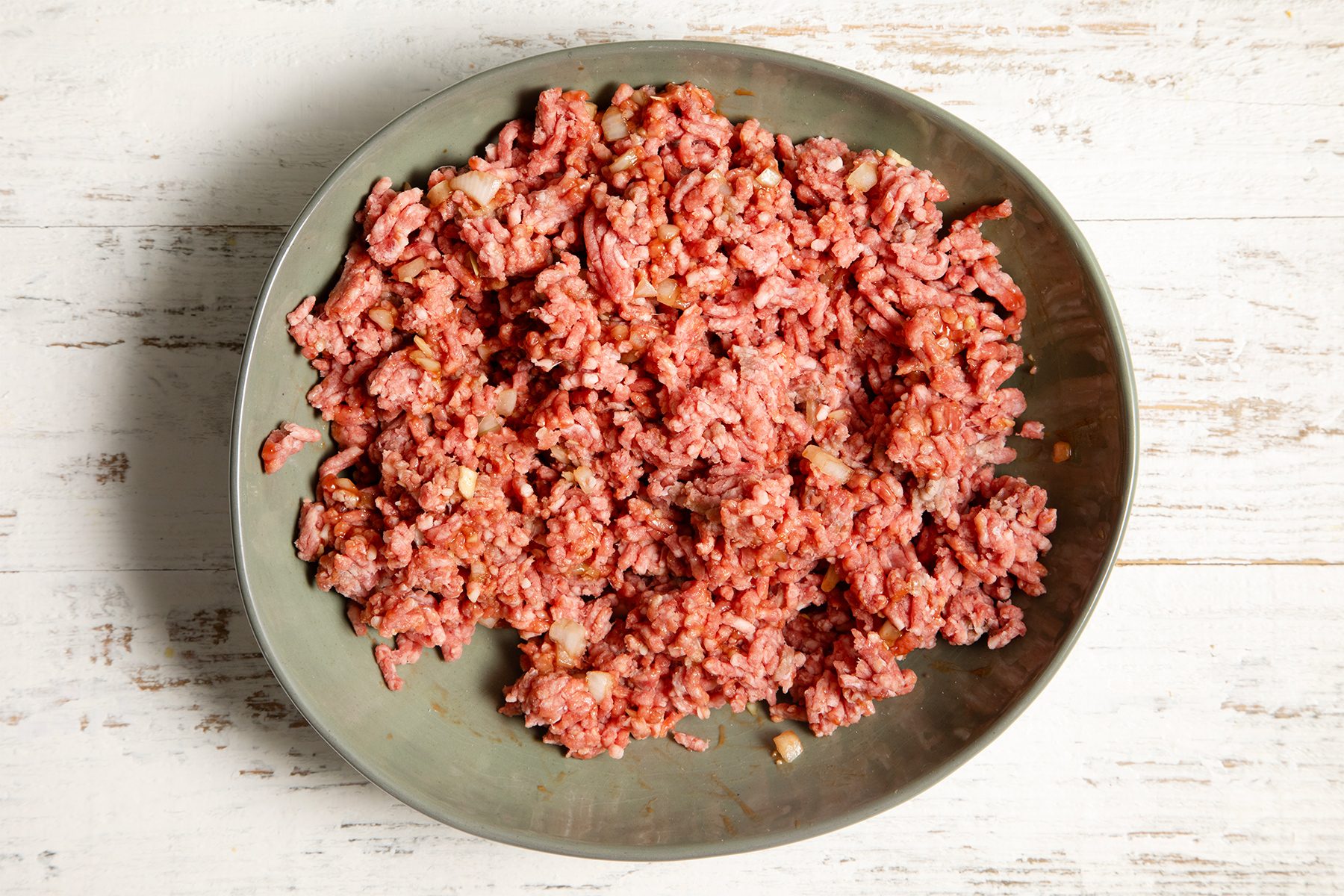 A large oval bowl filled with ground meat sits on a rustic white wooden surface. 