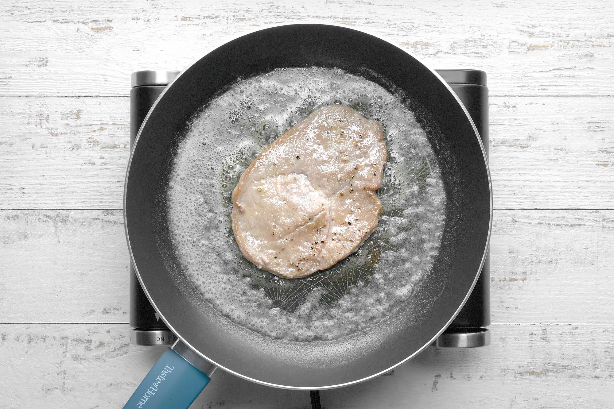 overhead shot; white wooden background; In a skillet cooking veal cutlet;