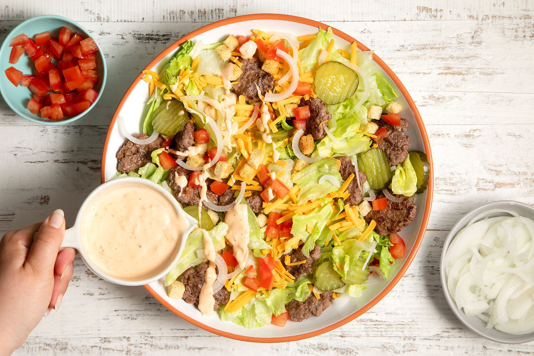 overhead shot; white textured background; Deluxe Cheeseburger Salad in a large bowl garnishing with salad dressing; tomatoes and sliced onion in small bowl;