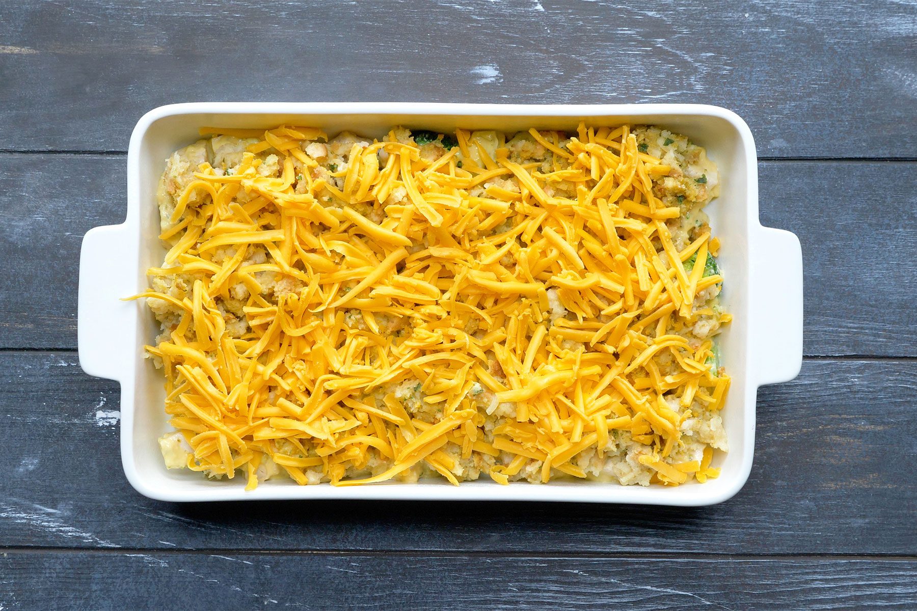 overhead shot; dark wooden background; Broccoli Chicken Casserole in baking dish topped with mixture and cheddar cheese;