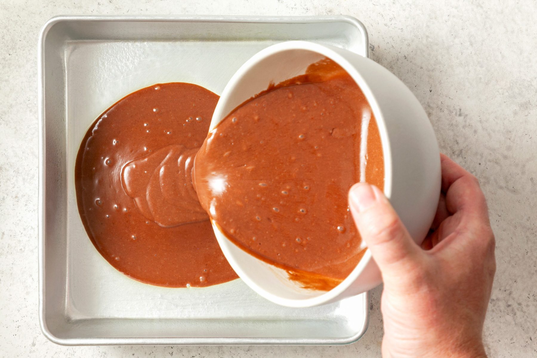 A hand pouring chocolate sauce into a baking pan