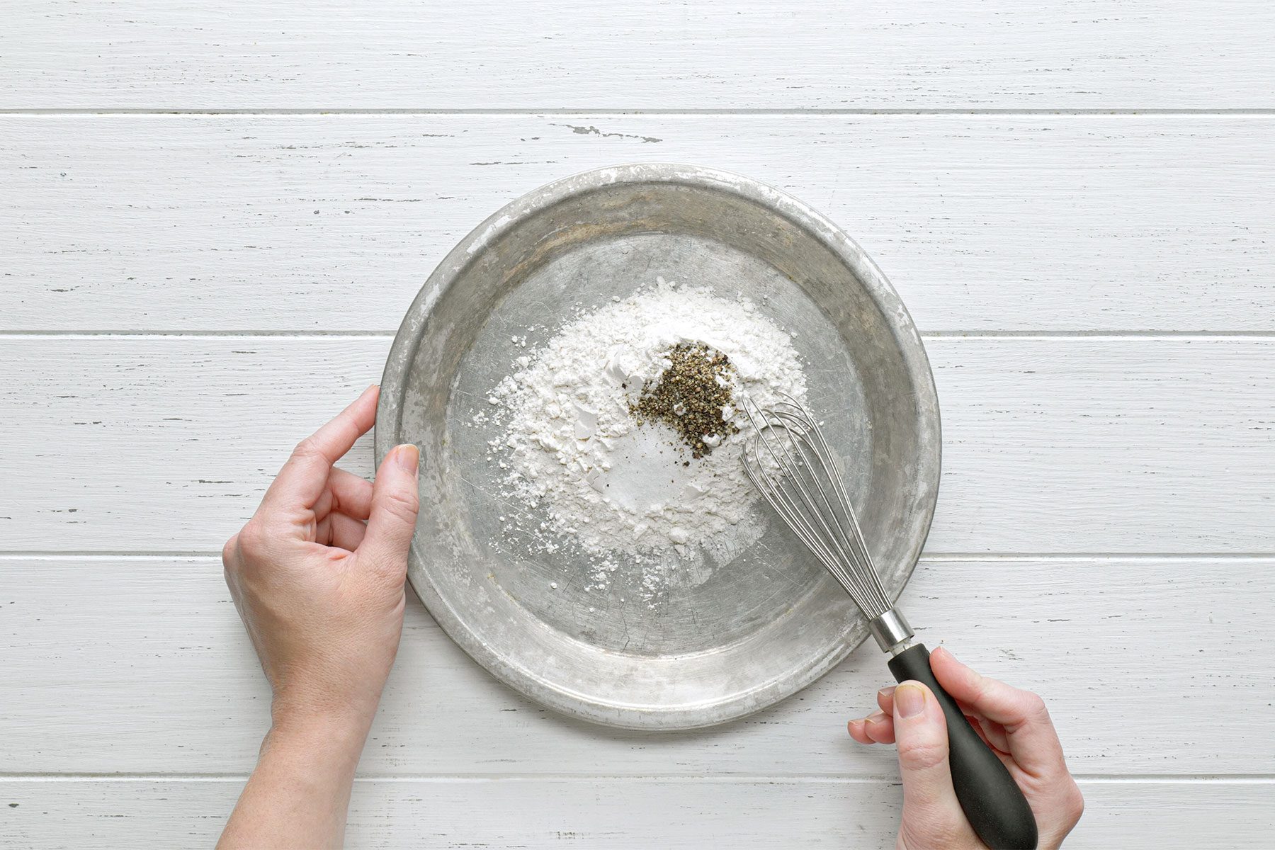 Overhead shot of a shallow bowl; mix flour salt and pepper; whisk tool; white wooden background;