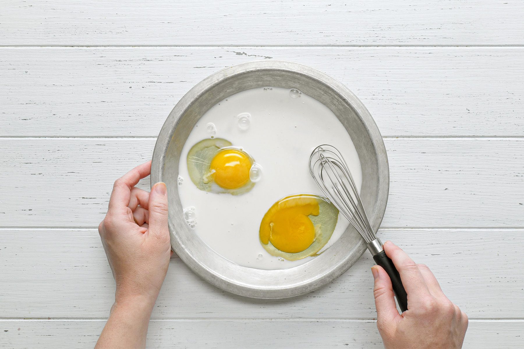 Overhead shot of whisk eggs and milk; in a separate shallow bowl; whisk tool; white wooden background;