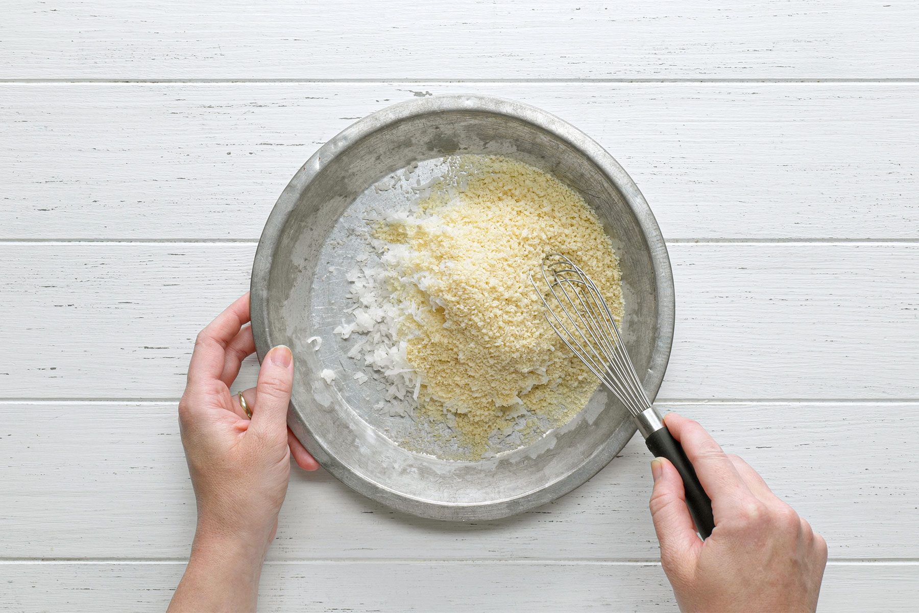 Overhead shot of combined coconut and bread crumbs in a third shallow bowl; whisk tool; white wooden background;