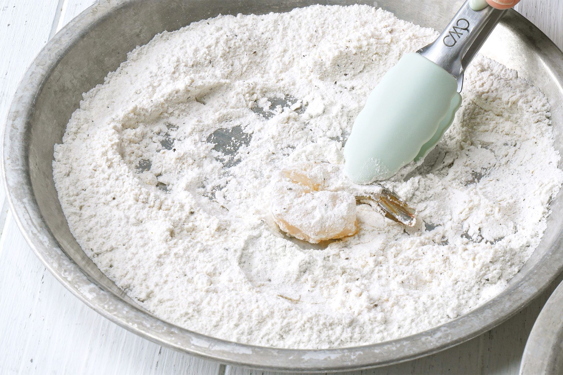 Close shot of dip shrimp in flour to coat both sides; shake off excess; with the help of tongs; white wooden background;