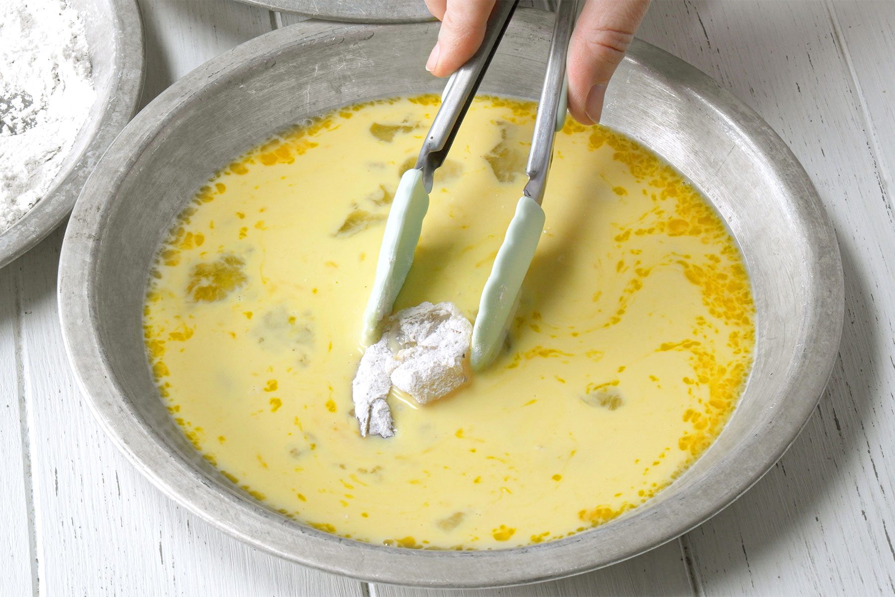 3/4 angle view shot of dip in egg mixture; with the help of tongs; white wooden background;