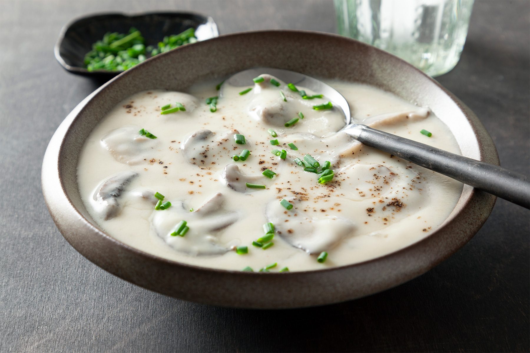 3/4 angle view shot of Quick Cream of Mushroom; served in large bowl; top with chives; spoon; dark texture background;