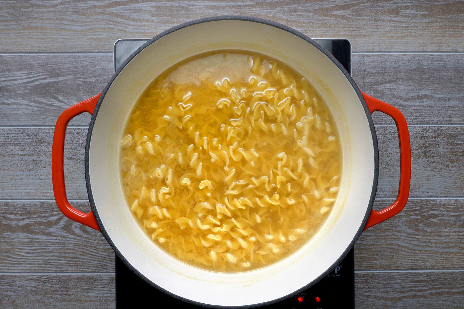 Overheda shot of a large saucepan; bring water and bouillon to boil; add noodles; cook uncovered until tender; induction; light brown wooden background;