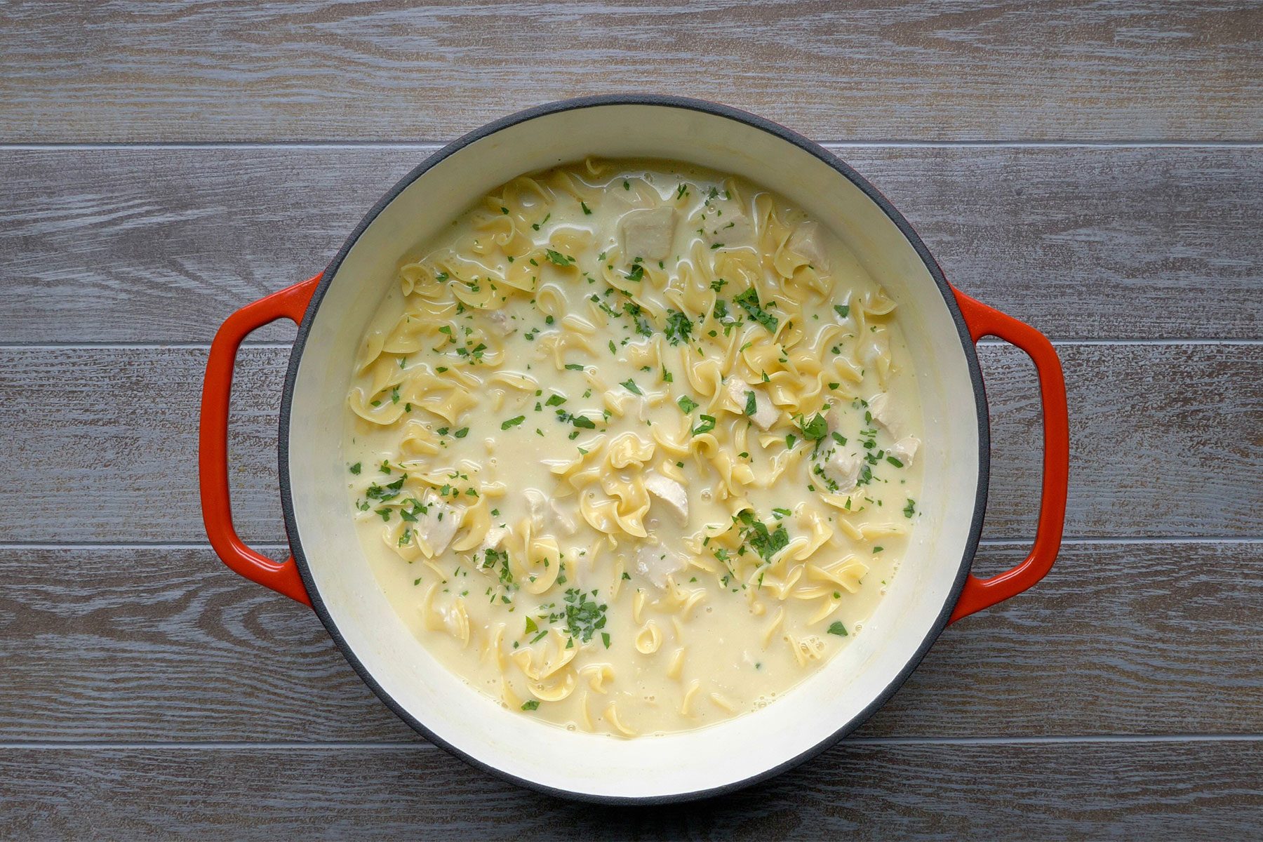 Overhead shot of Comforting Chicken Noodle Soup; sprinkled with minced parsley; light brown wooden background;