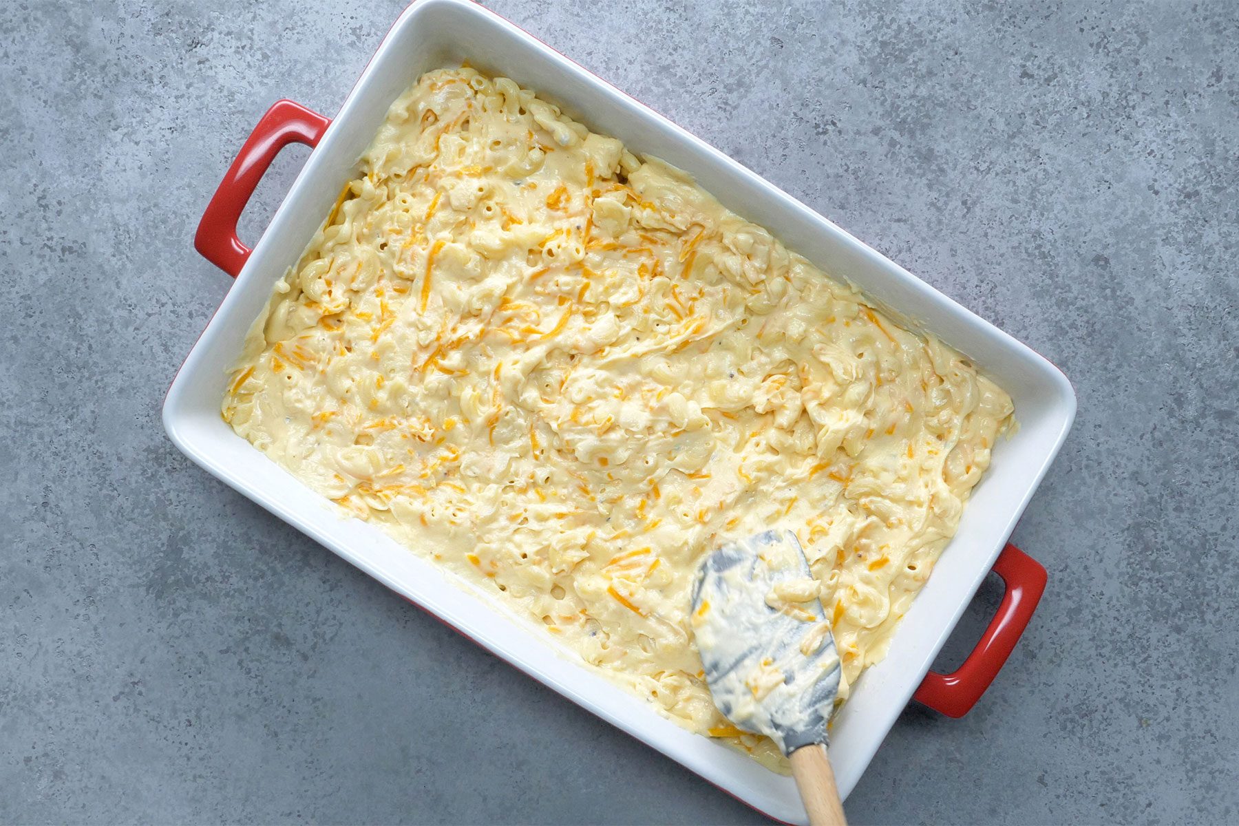 OVerhead shot of transferring the tossed macaroni and cream sauce to a greased baking dish; spatula; grey marble texture background;