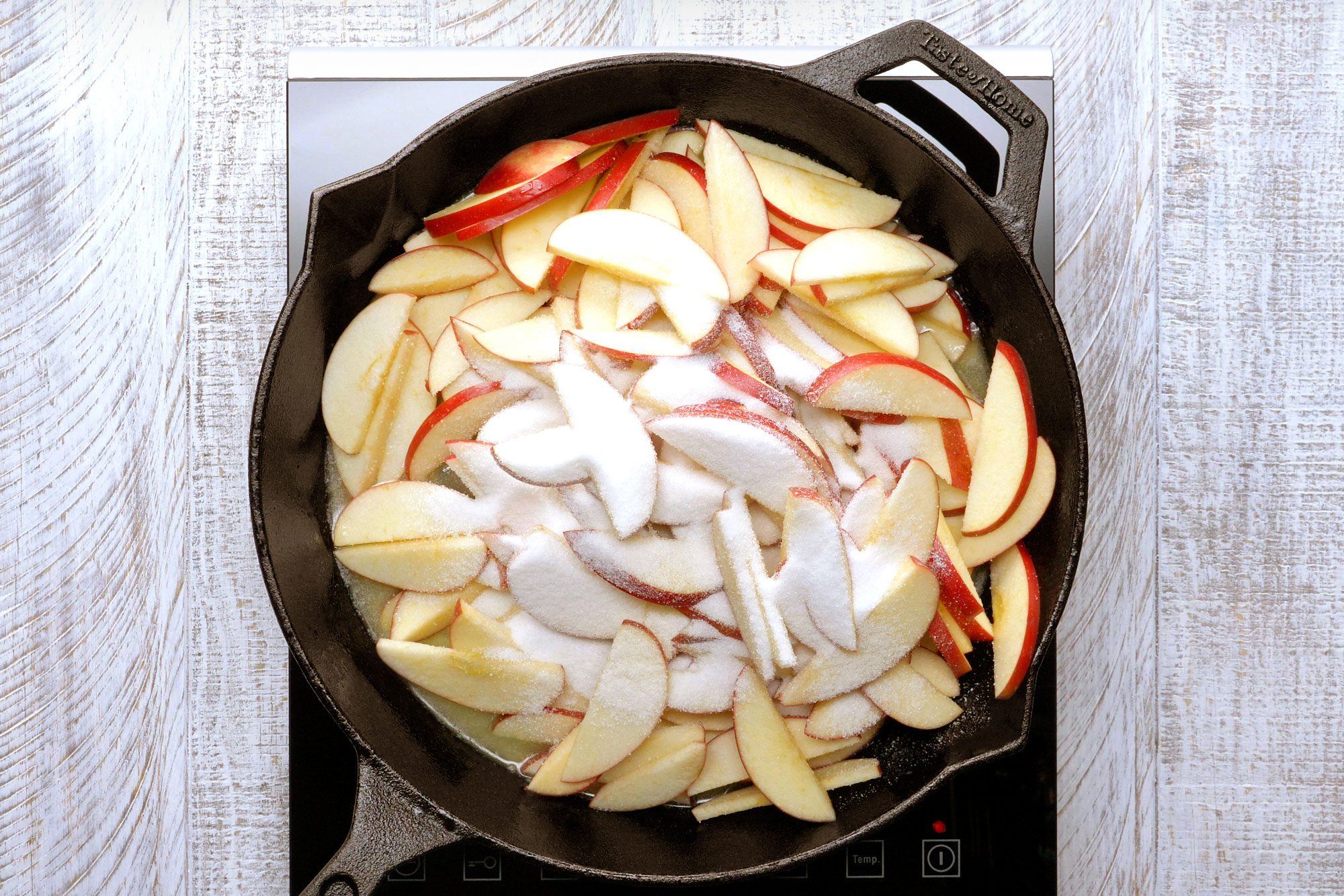 Overhead shot of apple slices, melted butter and 1/2 cup sugar in a large cast-iron skillet on wooden background