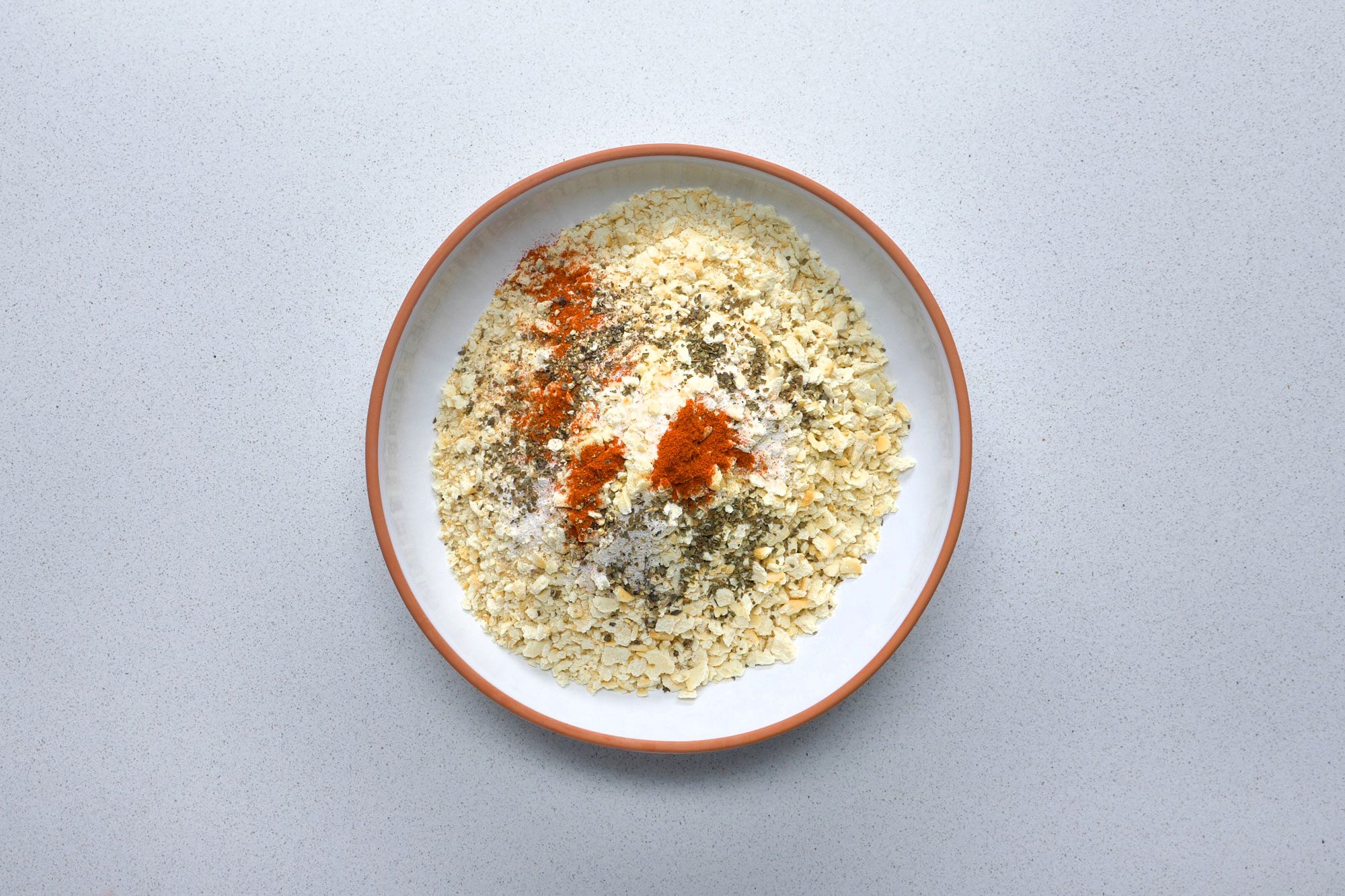 Overhead shot of crushed saltines, garlic salt, dried basil, paprika and pepper in a bowl on grey marble background