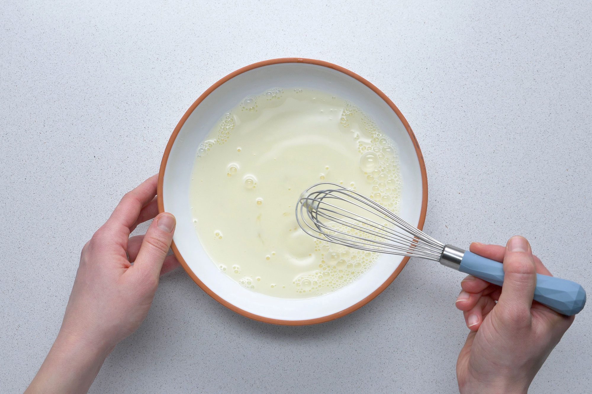 Overhead shot of beating egg and milk in a bowl using whisk on grey marble background
