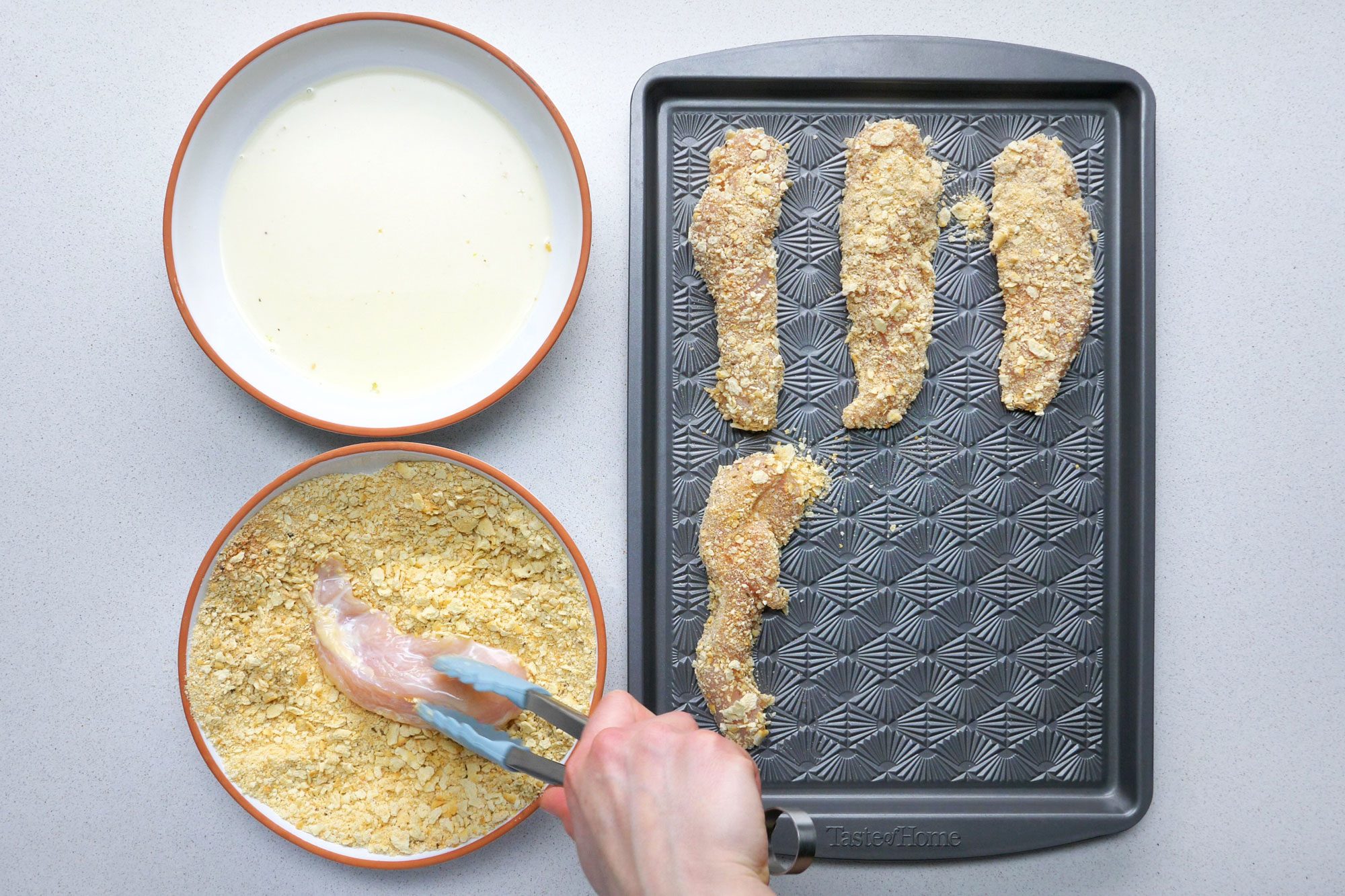 Overhead shot of dipping chicken into egg mixture; then cracker mixture; keeping it on side tray using tongs; grey marble background;