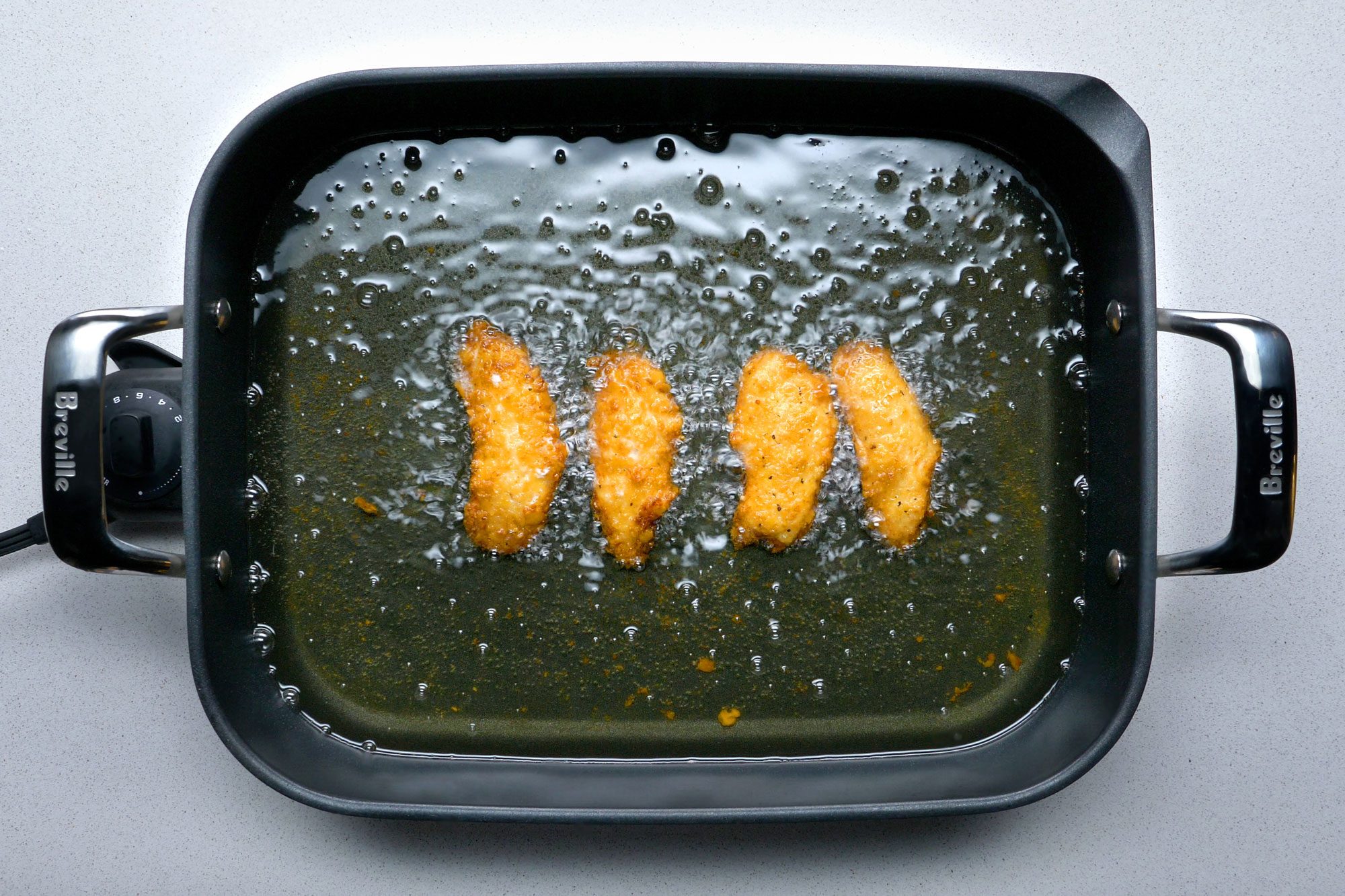 Overhead shot of deep-fat frying chicken strips in an electric skillet on grey marble background