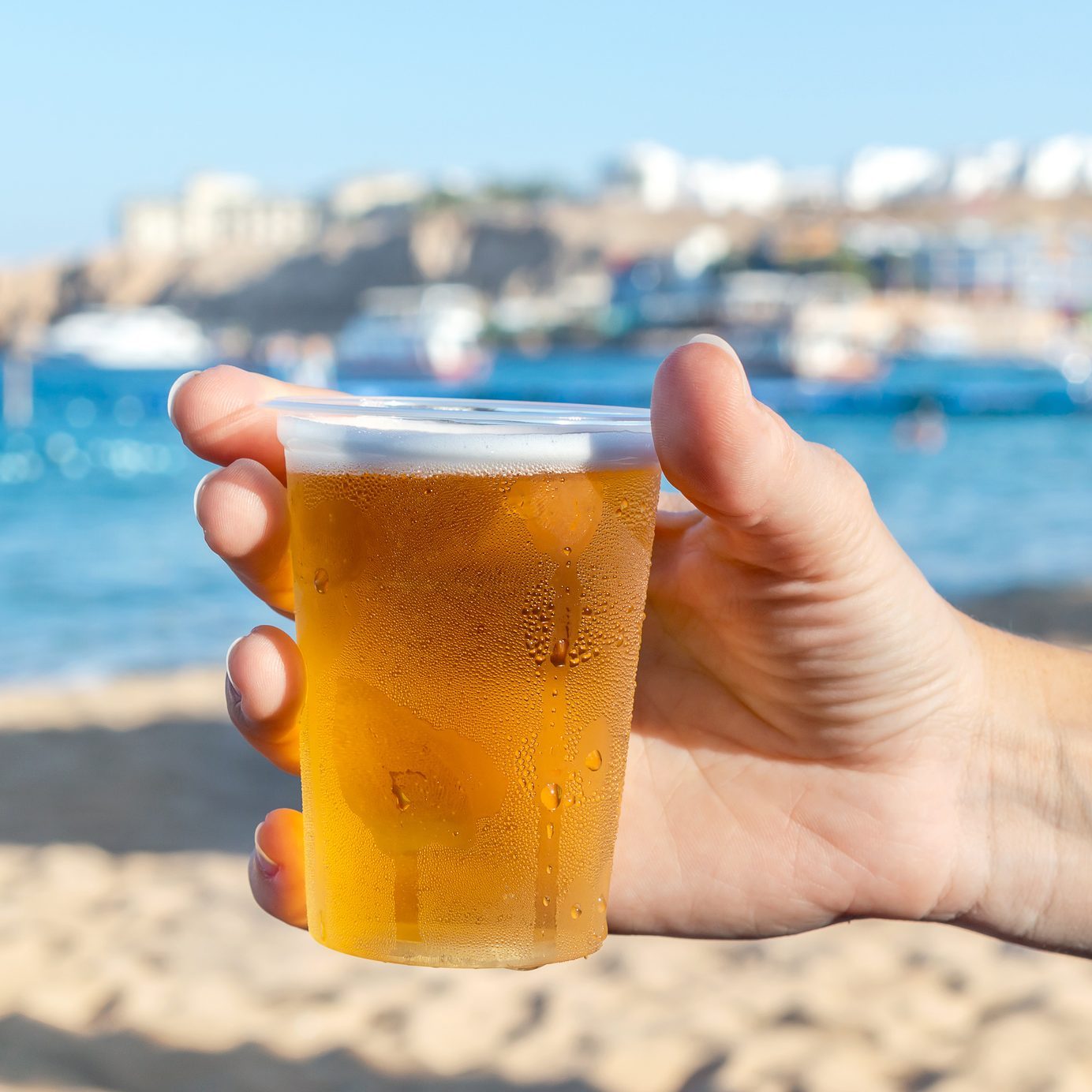 Woman's hand holding plastic glass of beer on sea beach.
