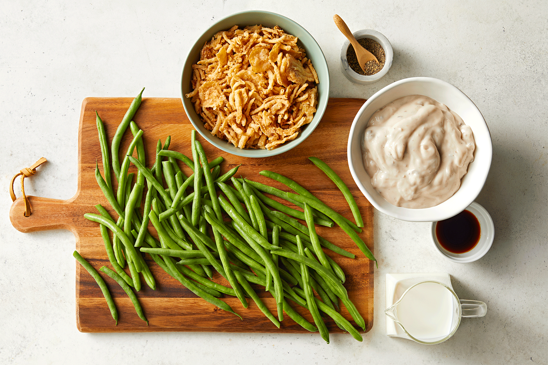 A wooden cutting board with fresh green beans and bowls of fried onions, creamy sauce, milk, soy sauce, and black pepper arranged on a white surface. 