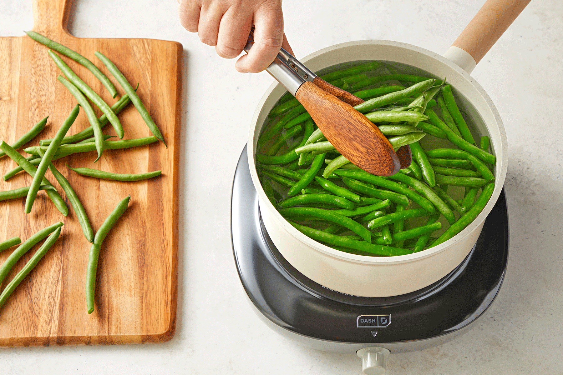 A person uses tongs to place green beans into a pot of boiling water on an induction cooktop. 