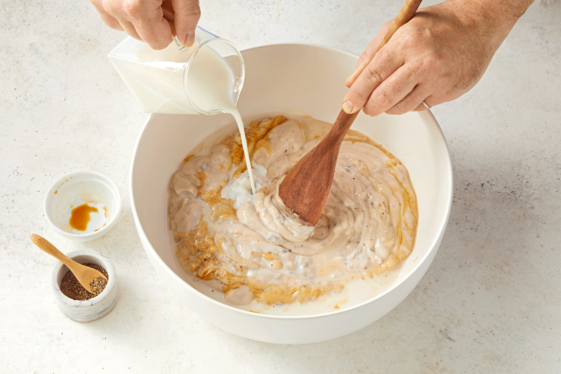 A person is pouring milk from a small pitcher into a large, white mixing bowl while stirring with a wooden spoon.