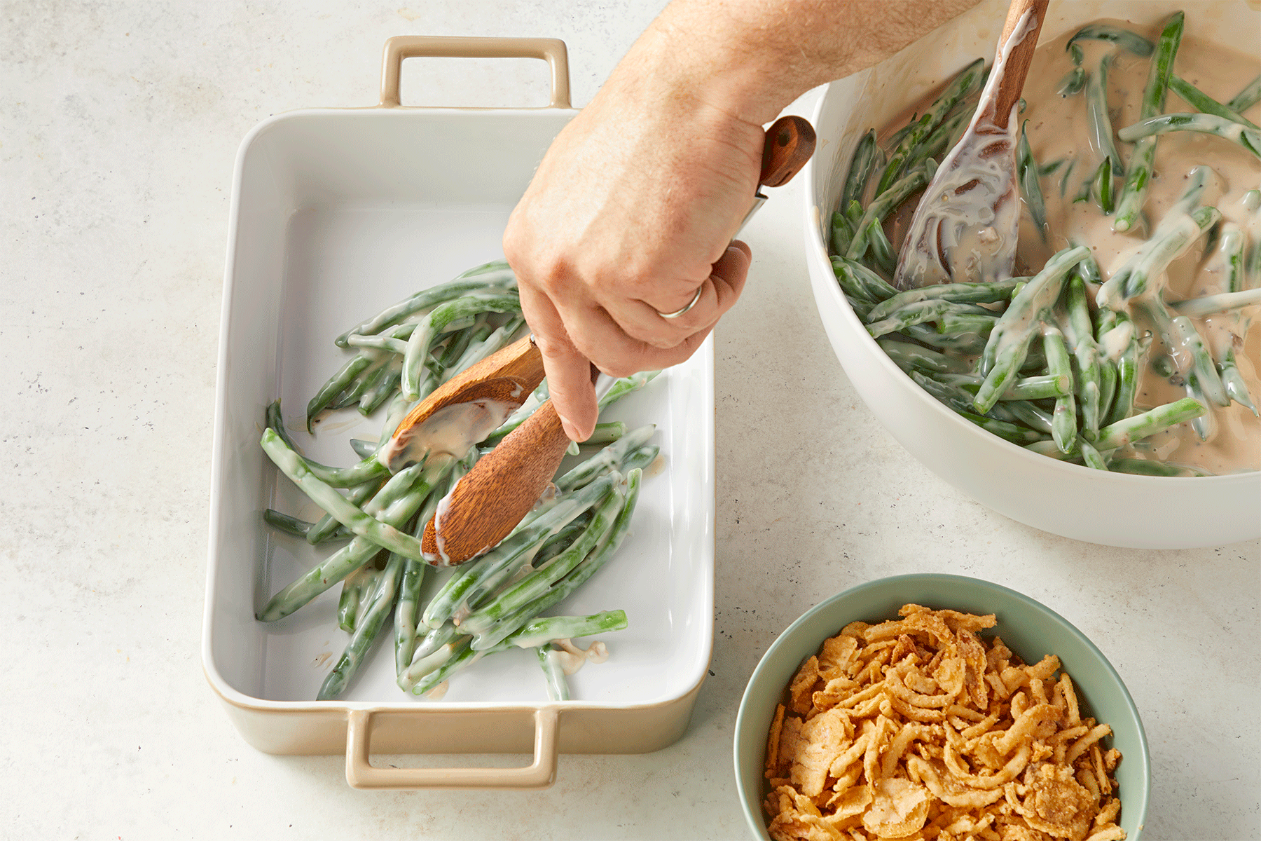 A person uses wooden tongs to transfer creamy green beans from a mixing bowl to a white rectangular baking dish. 