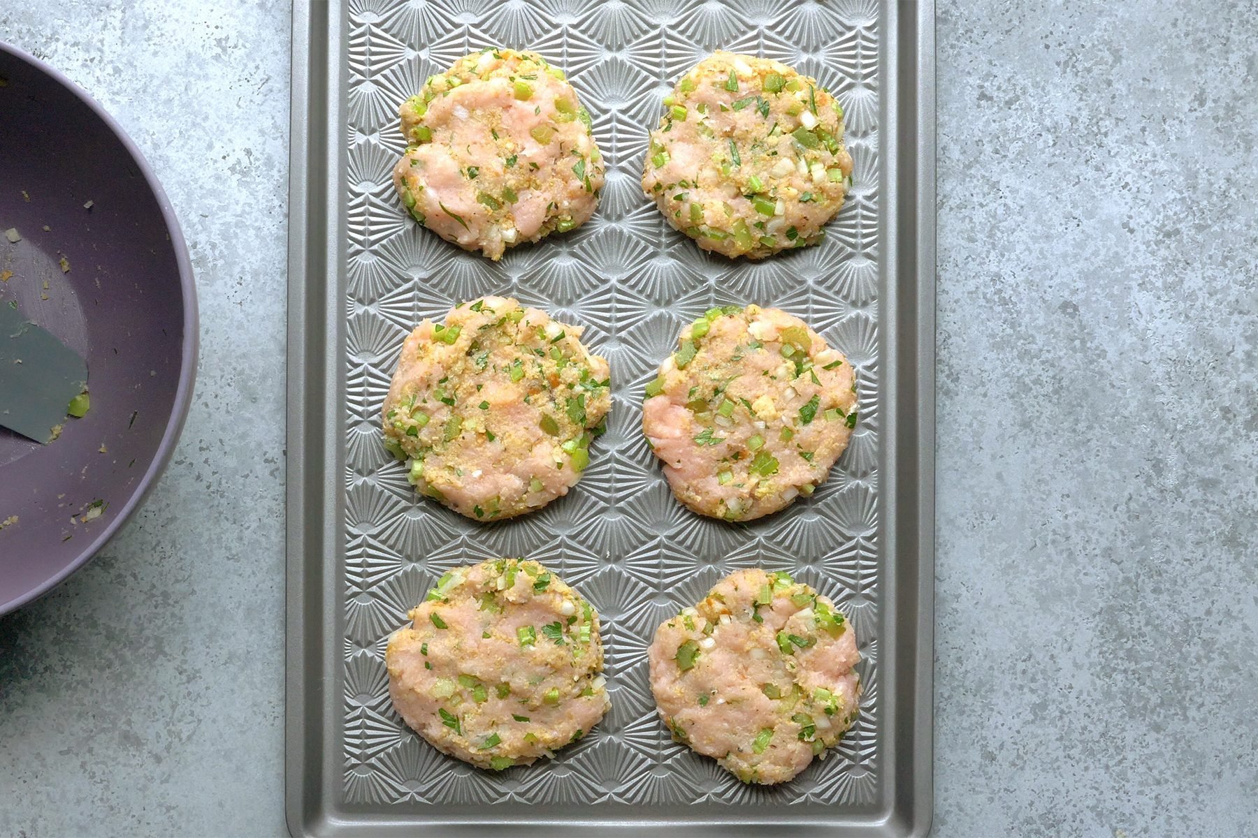 A baking sheet with six uncooked chicken patties embedded with chopped green vegetables.