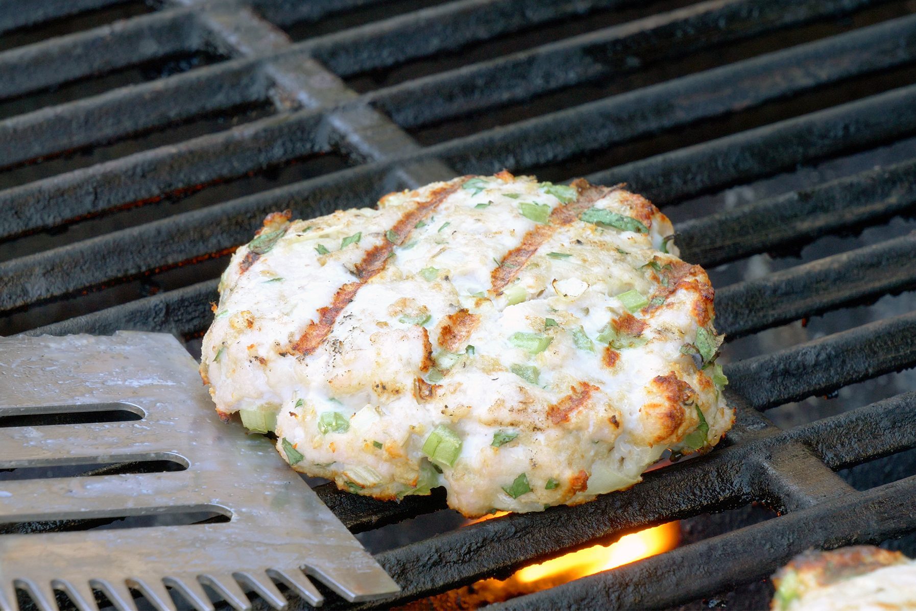 A close-up of a chicken patty mixed with chopped green onions being grilled on an open flame barbecue.