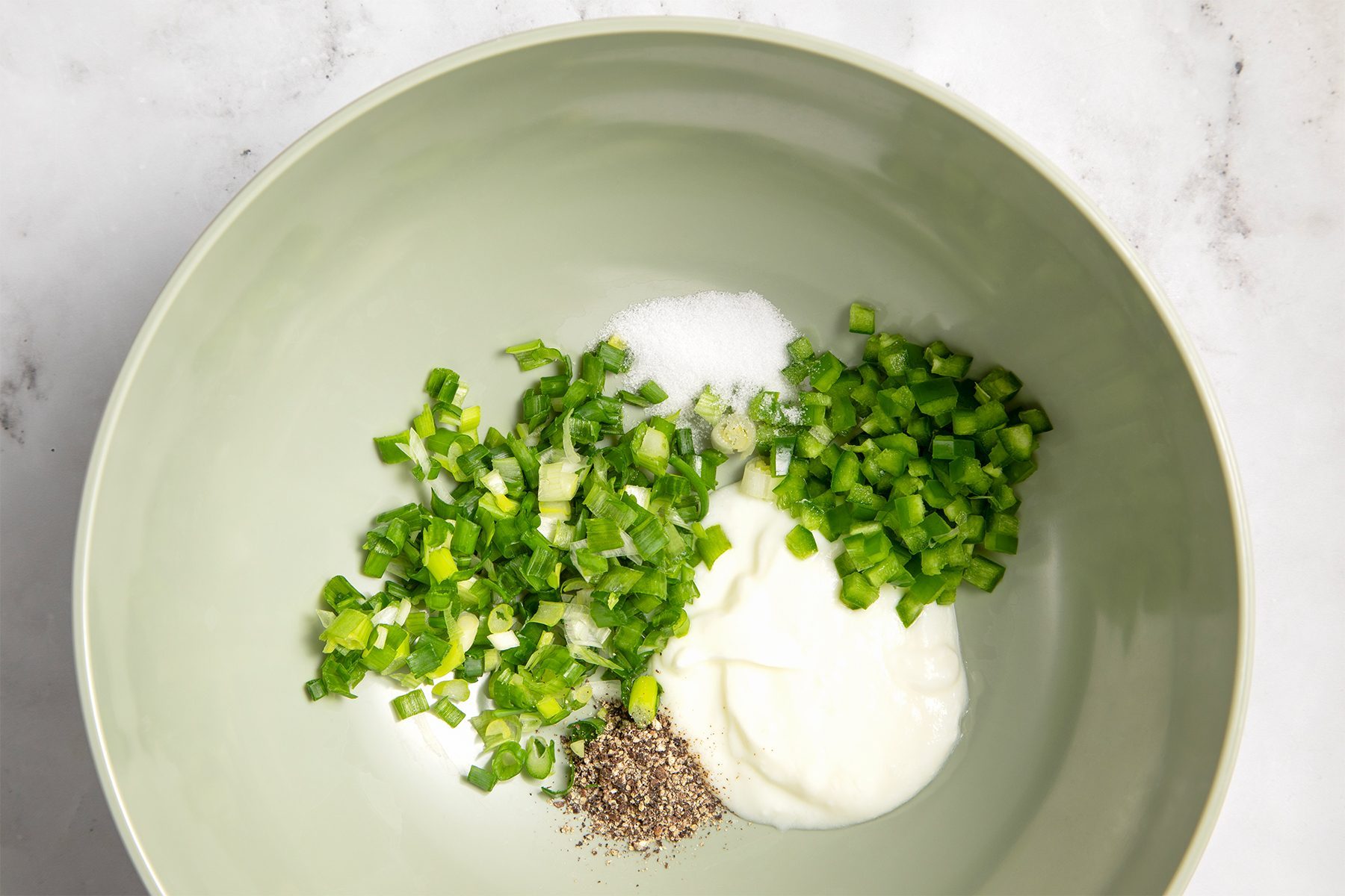 A light green bowl containing chopped green onions, diced green bell peppers, a small pile of sugar, a dollop of yogurt, and a sprinkle of ground black pepper, all neatly arranged on a light surface.
