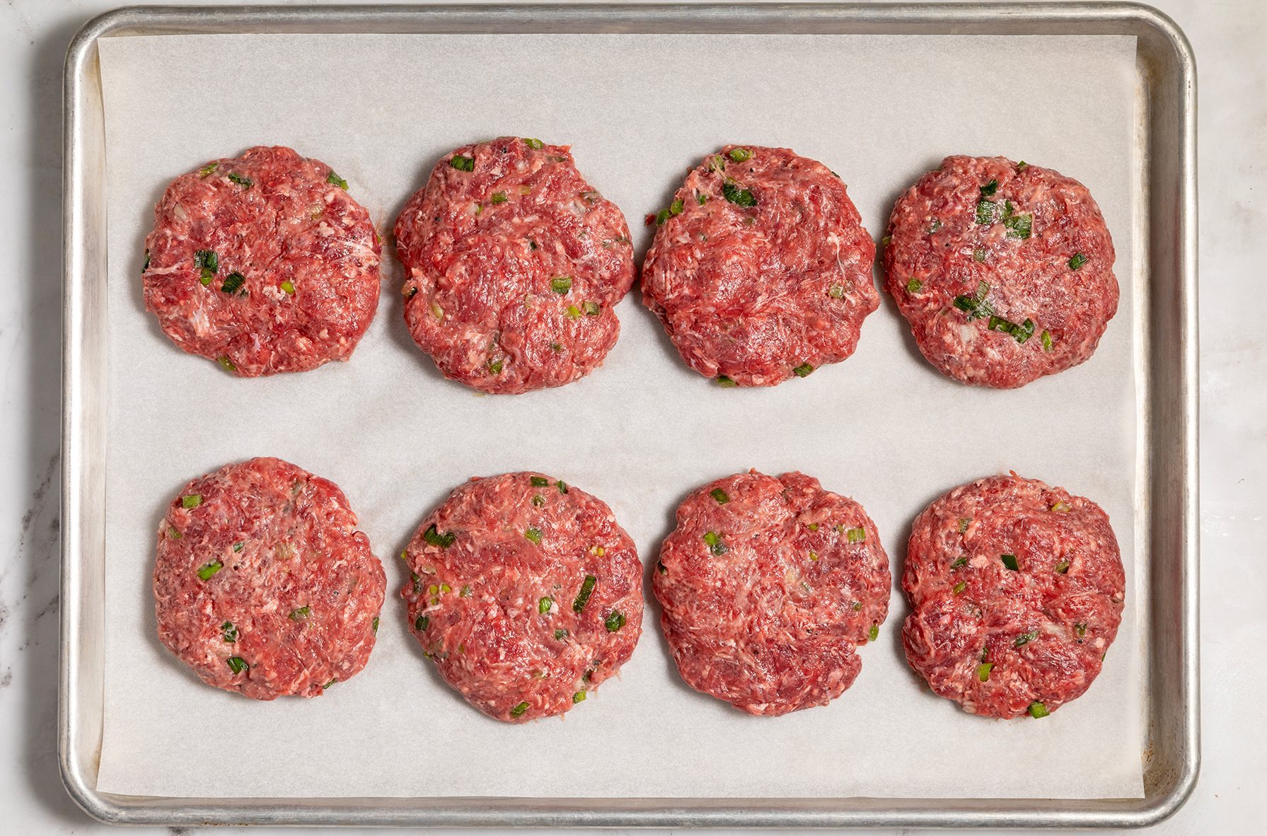Eight raw beef patties with green seasoning flecks are evenly spaced on a parchment-lined baking sheet. 