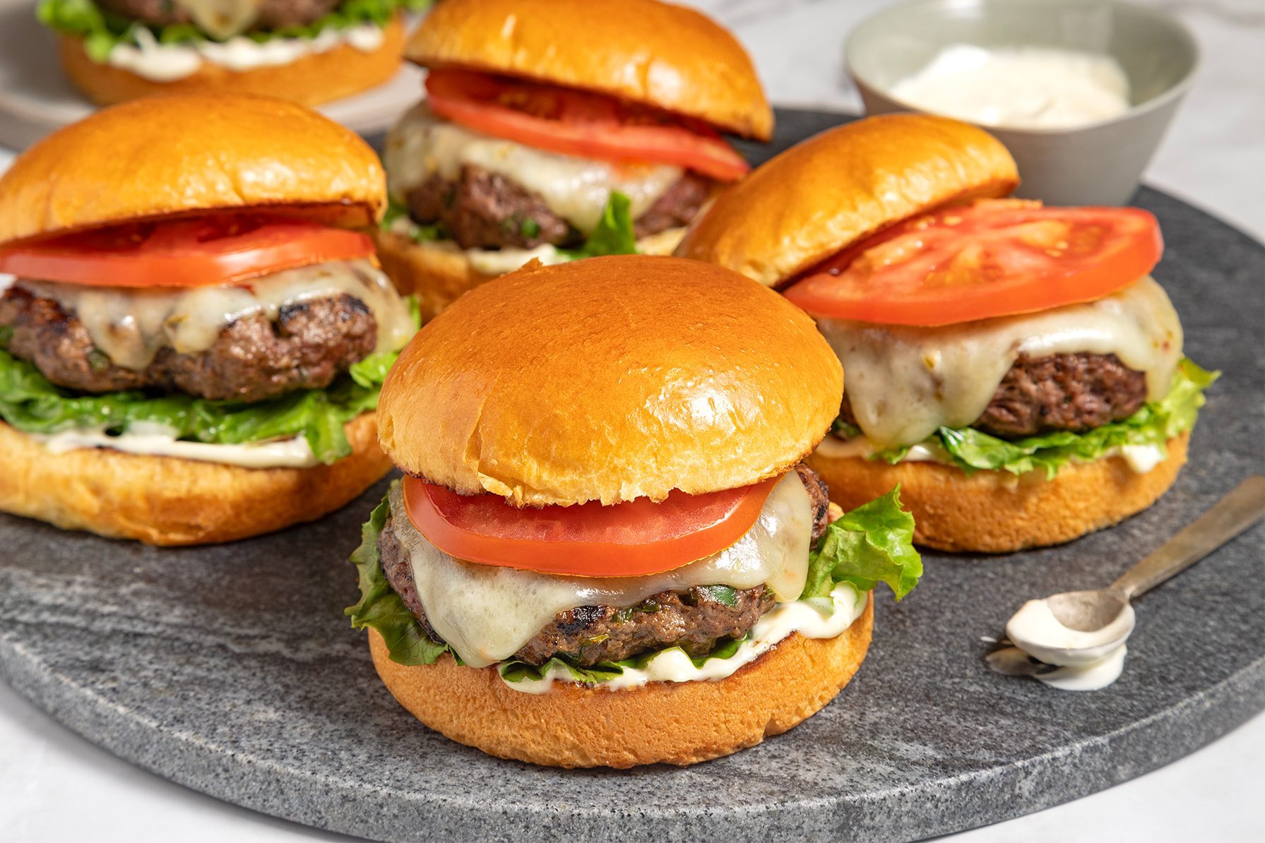 A plate of five cheeseburgers with melted cheese, lettuce, and tomato slices on a round, dark grey serving platter.