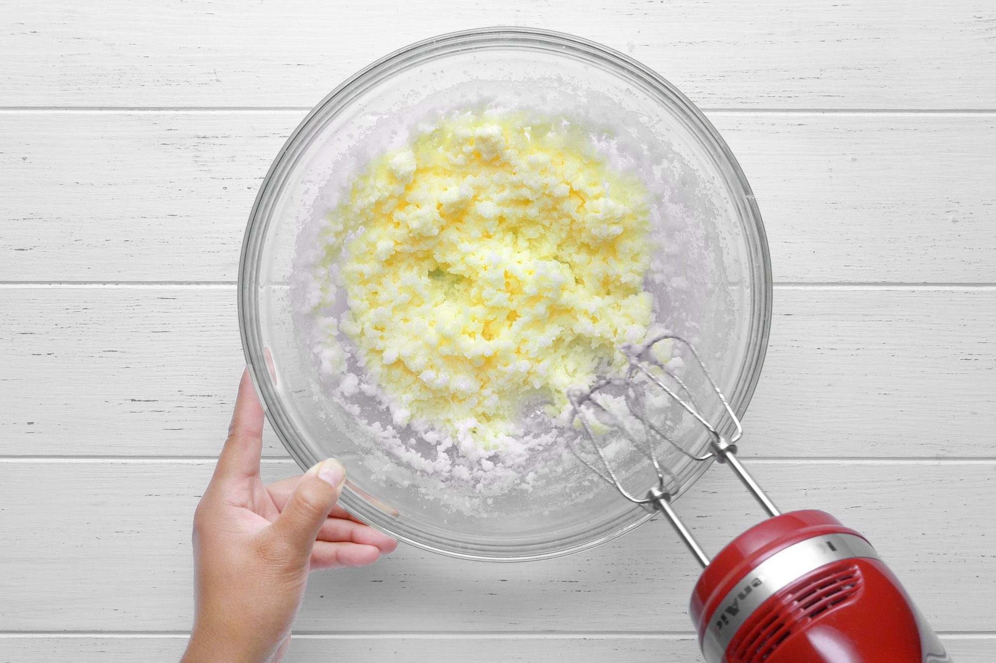 Overhead shot of blending granulated sugar, melted butter and oil using hand mixer on wooden surface