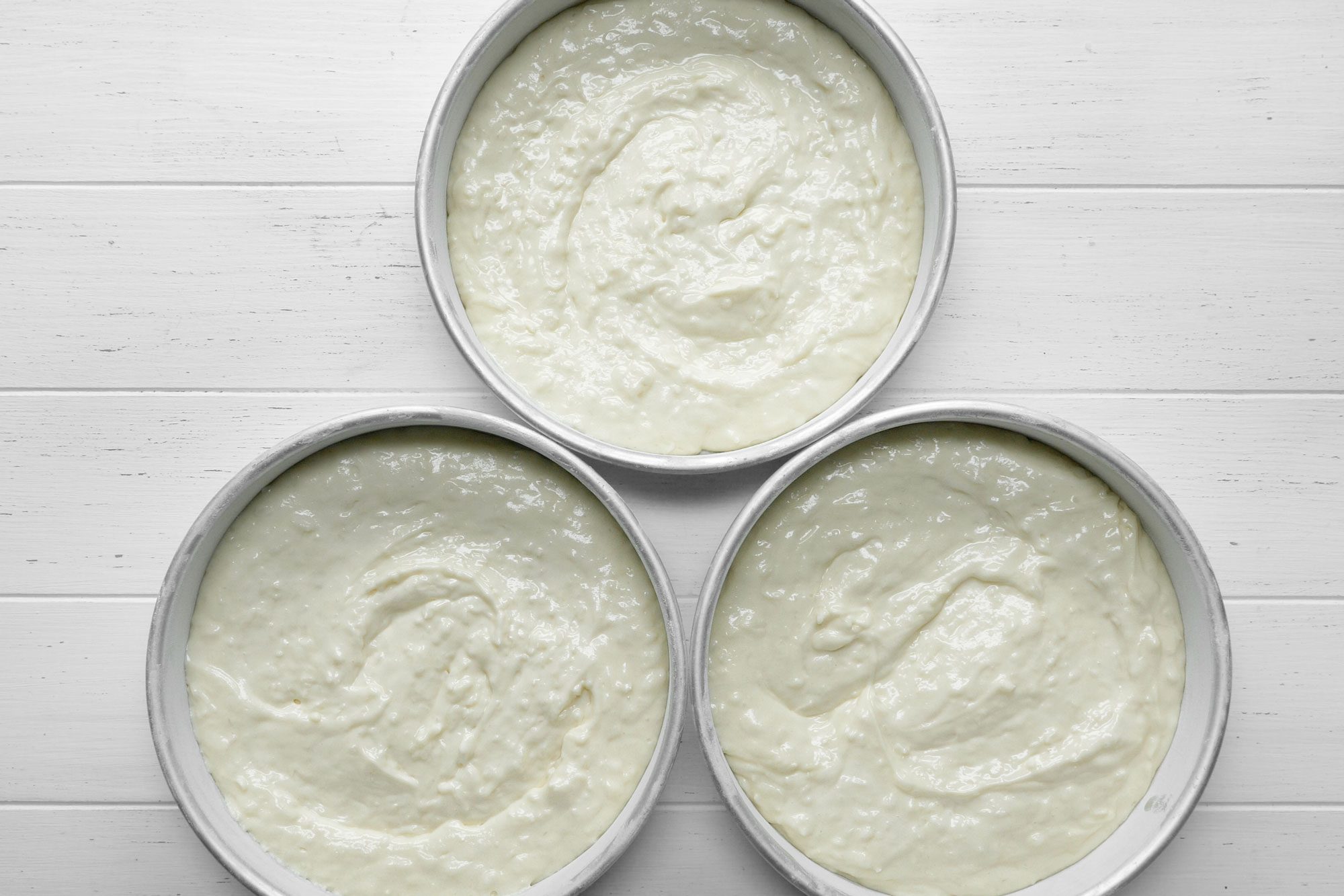 Overhead shot of batter in 3 greased round baking pans; white wooden background;