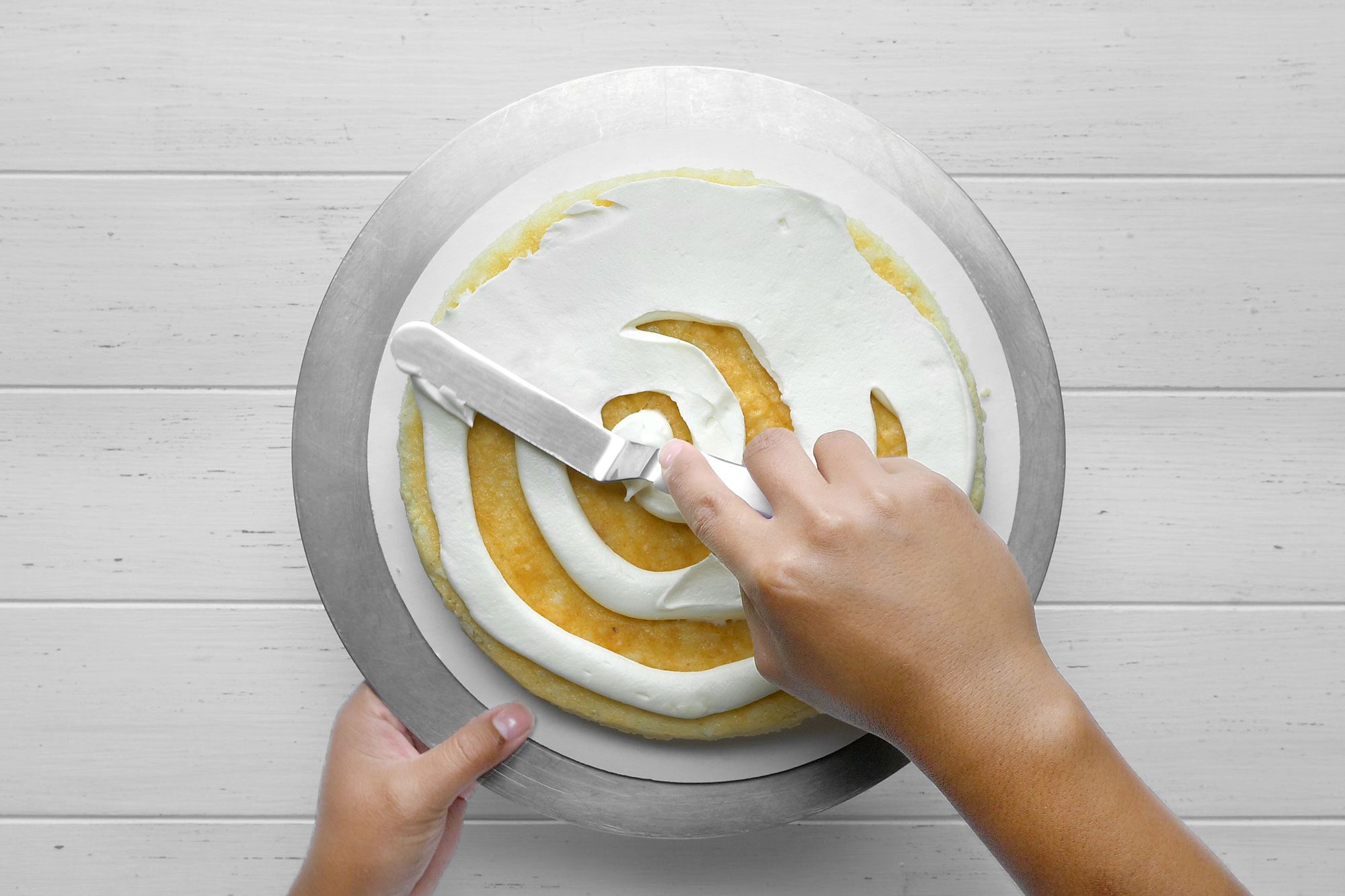 Overhead shot of place cake layer on a serving plate; spreading with 1/2 cup frosting; white wooden background;