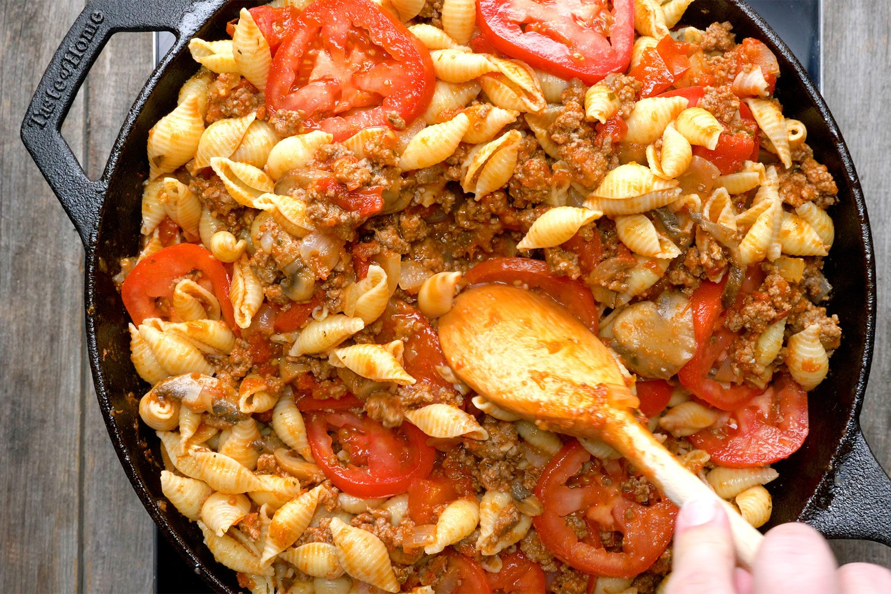 Overhead shot of large skillet; gently stir in sliced plum tomatoes; induction stove; wooden background;