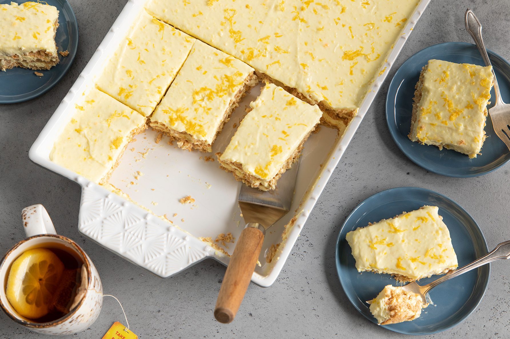 A white rectangular baking dish contains lemon squares, with several pieces already cut. Three lemon squares are placed on blue plates, each with a fork.
