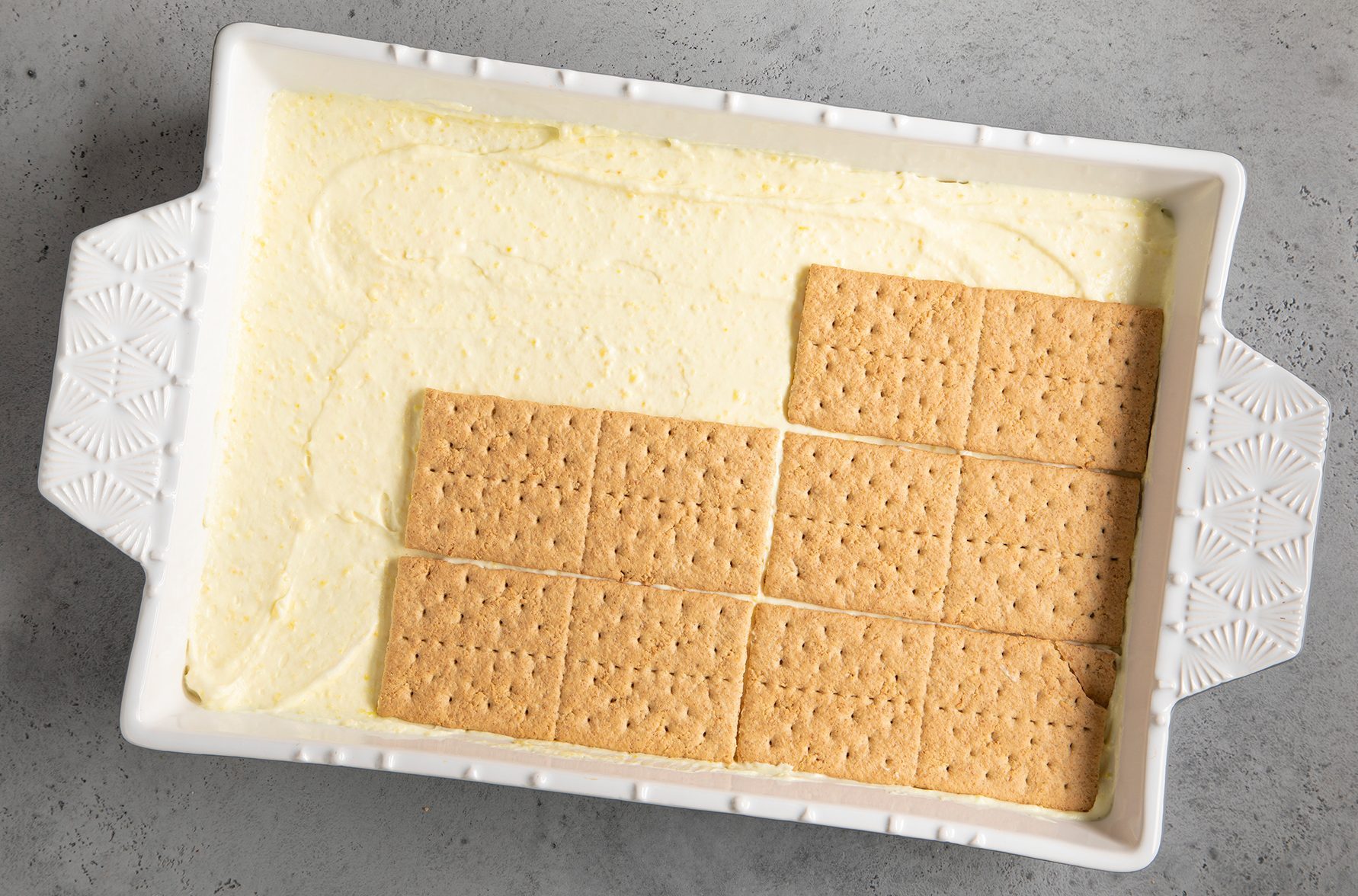 A white rectangular baking dish contains a layer of creamy mixture spread evenly, with rectangular graham crackers placed on top, covering about two-thirds of the dish.