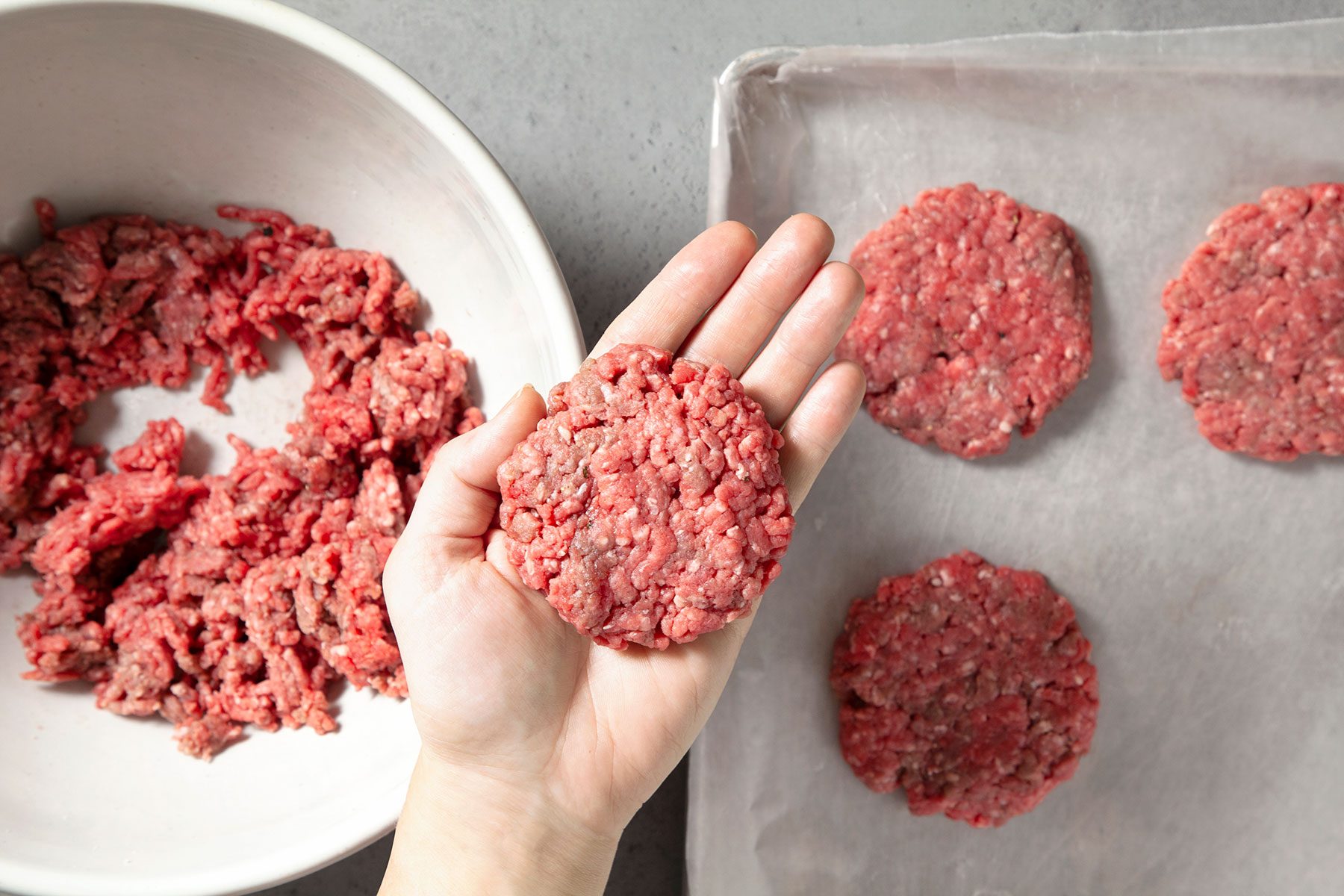 Overhead shot of large bowl; combine beef; salt and pepper; mix well; shape into thick patties; baking tray with sheet; grey marble background;