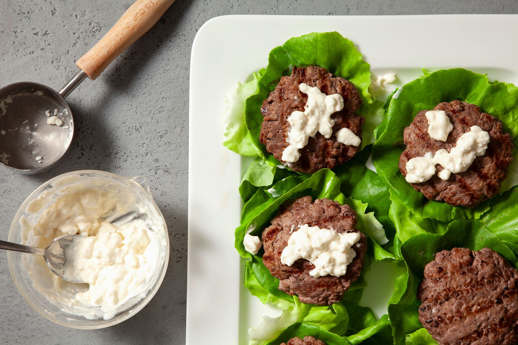 Overhead shot of combine feta and miracle whip; spread over burgers; white tray; spoon; grey marble background;