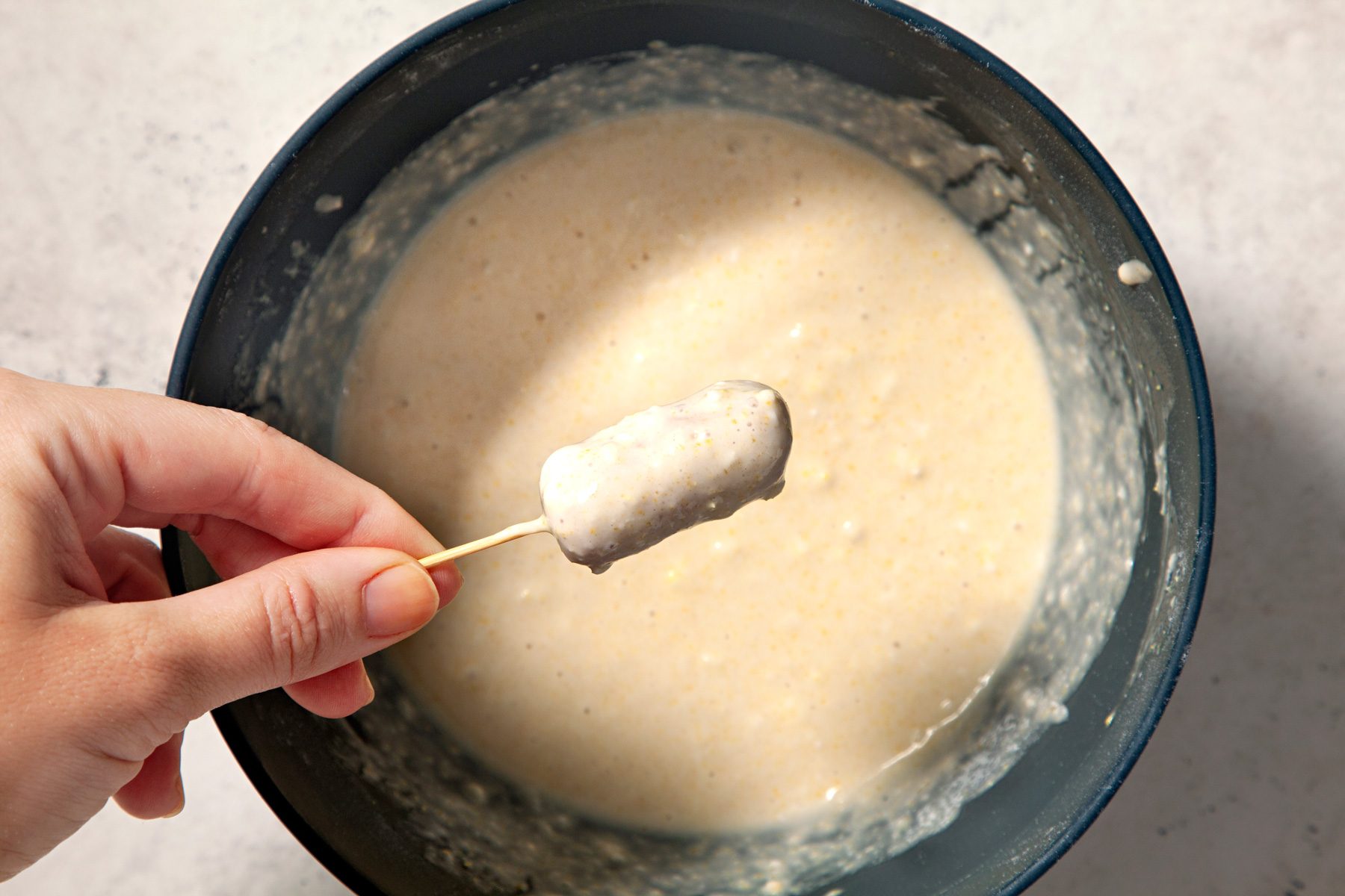 A hand is dipping sausage into batter