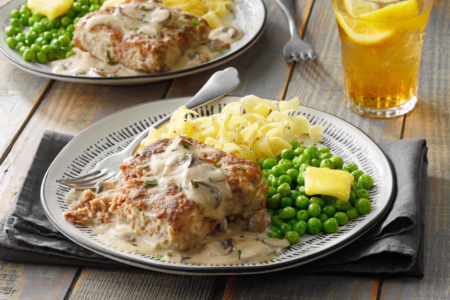 Two plates of hearty meals with sliced pork chops covered in creamy mushroom sauce, sides of green peas, and buttered egg noodles. Each plate is garnished neatly. In the background is an ice-cold drink with lemon slices. The meal is set on a wooden table.