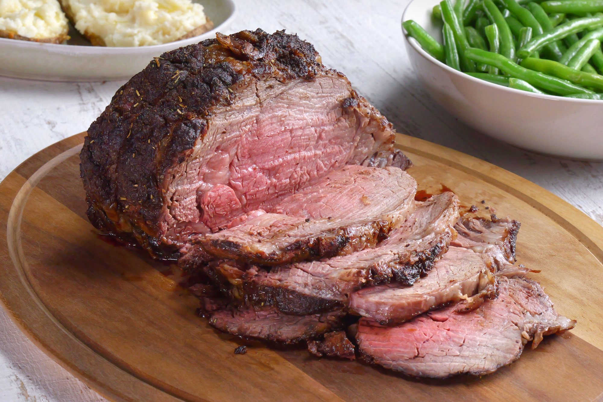 3/4 angle view shot of Seasoned Ribeye Roast; serve on large wooden platter; with fresh beans and mashed potatoes; white wooden background;